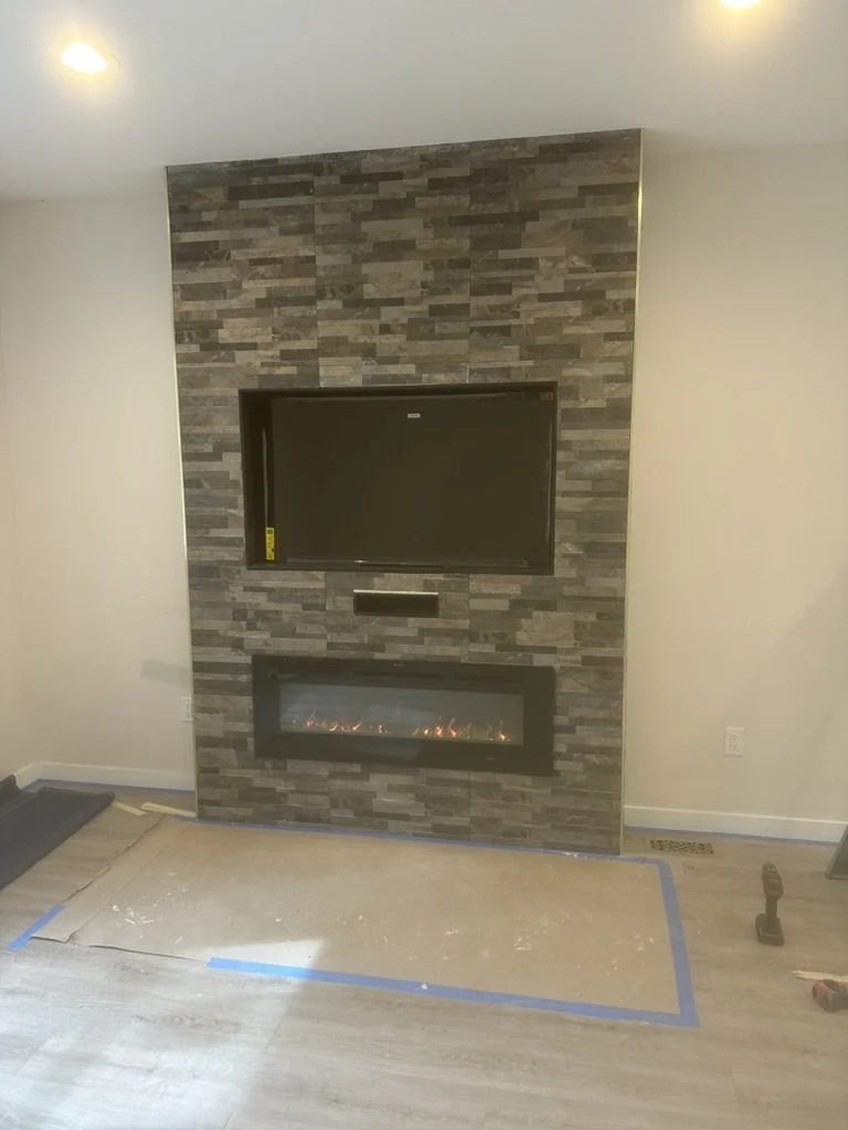 Living room with a stone accent wall, mounted flat-screen TV, and electric fireplace below. The floor appears to be light-colored wood or laminate, with protective paper and blue tape on the floor, and some tools nearby.