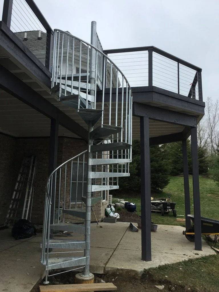 Metal spiral staircase leading from a lower patio area to an upper deck with black railing, attached to a house exterior.