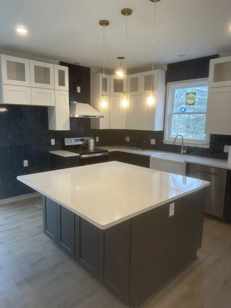 Modern kitchen with white upper cabinets, black lower cabinets, a central island with a white countertop, black walls, a window above the sink, and three hanging pendant lights.