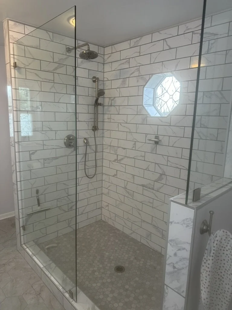 A walk-in shower with clear glass walls, tiled with white marble subway tiles, featuring a round hexagonal window, a rainfall showerhead, and a handheld showerhead.