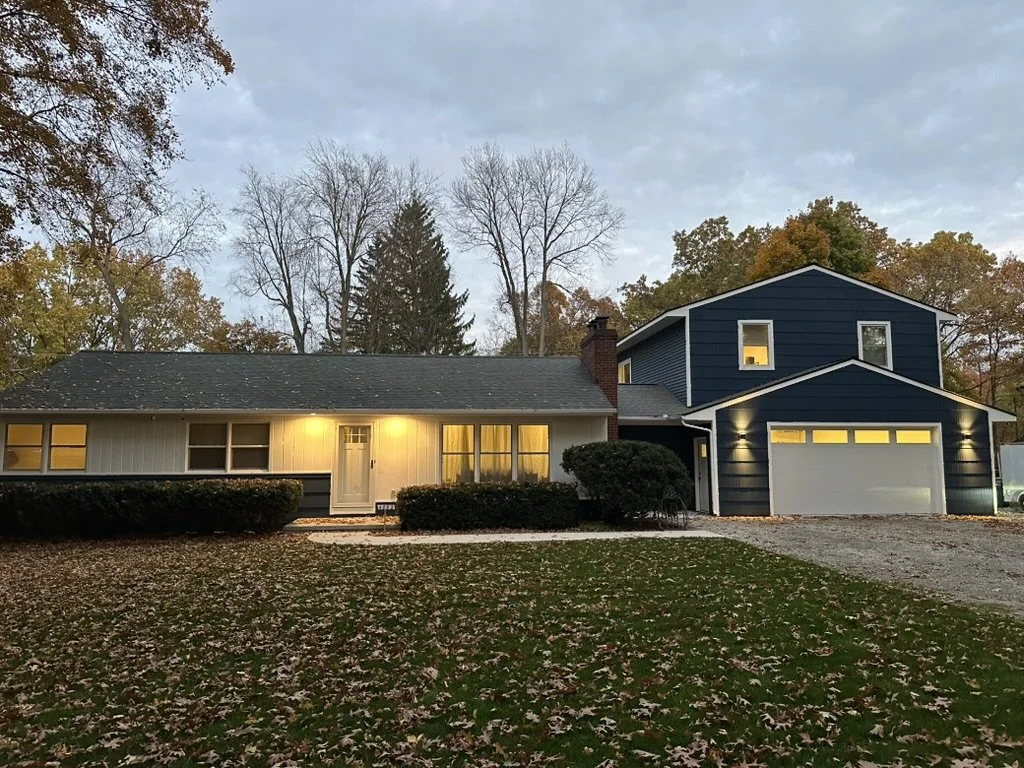 A two-story house with a dark blue and white exterior, illuminated windows, a front door, and a two-car garage, situated in a yard with fallen leaves and a driveway, surrounded by trees with some autumn foliage.