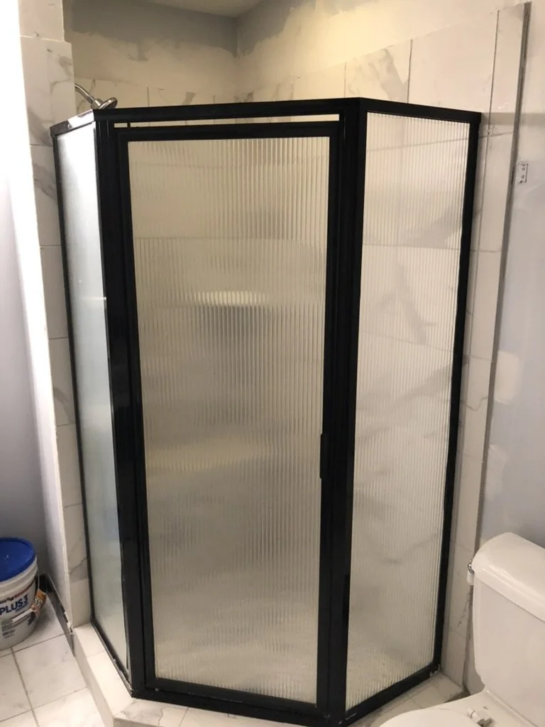 A corner shower with textured frosted glass doors and black frame, located next to a toilet and a bucket in a bathroom with marble walls.