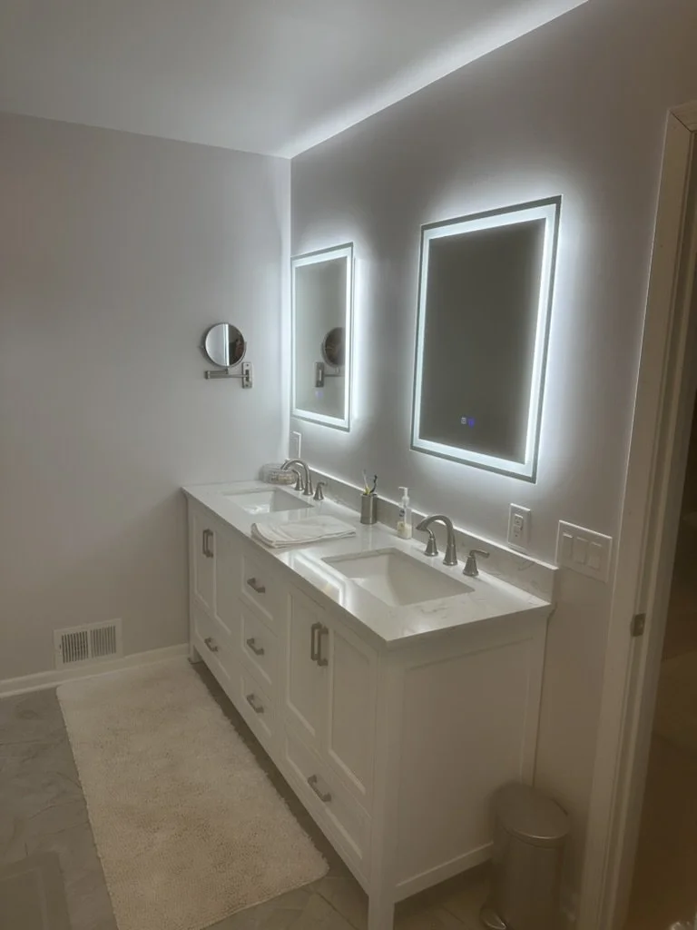 A modern bathroom with two illuminated mirrors above a white double vanity, featuring two sinks and silver fixtures. There are small wall-mounted magnifying mirrors, a towel on the countertop, soap dispenser, and a trash can. The floor has gray tiles