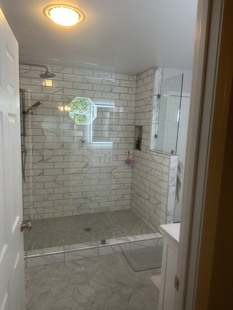 Bathroom shower area with white subway tiles, a small window with an octagonal shape, a rain shower head, and a handheld shower, with a small built-in niche for toiletries.
