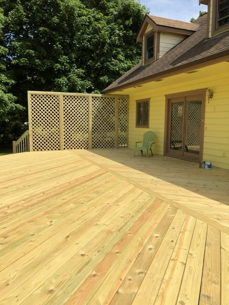 Newly built wooden deck attached to yellow house with sliding glass doors, a green chair, and lattice fencing in a backyard with lush green trees.