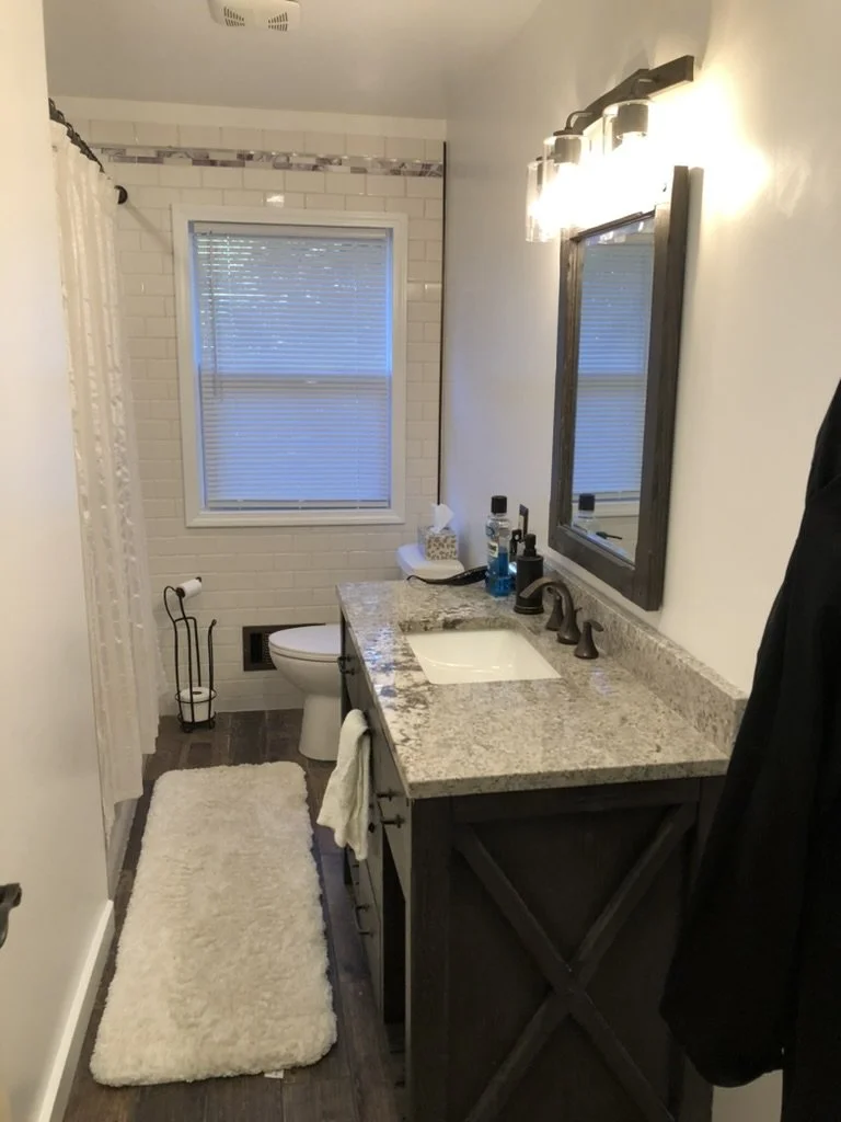 Bathroom with white brick wall, window with closed blinds, granite countertop vanity with dark wood cabinet, mirror, and bright light fixture, toilet, shower curtain, and various toiletries on the counter, and a white rug on dark wood floor.
