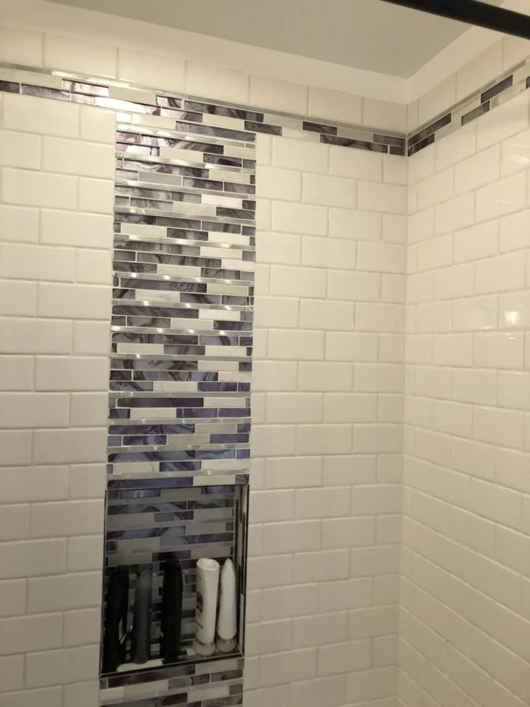 A shower wall with white subway tiles and a decorative vertical strip of gray and white mosaic tiles. There is a built-in niche holding shampoo and conditioner bottles.