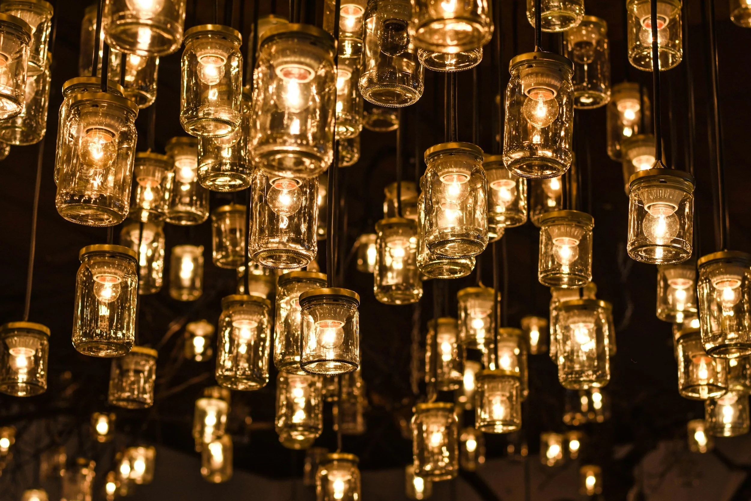 Multiple hanging glass jars with lit light bulbs inside, creating a warm yellow glow against a dark background.