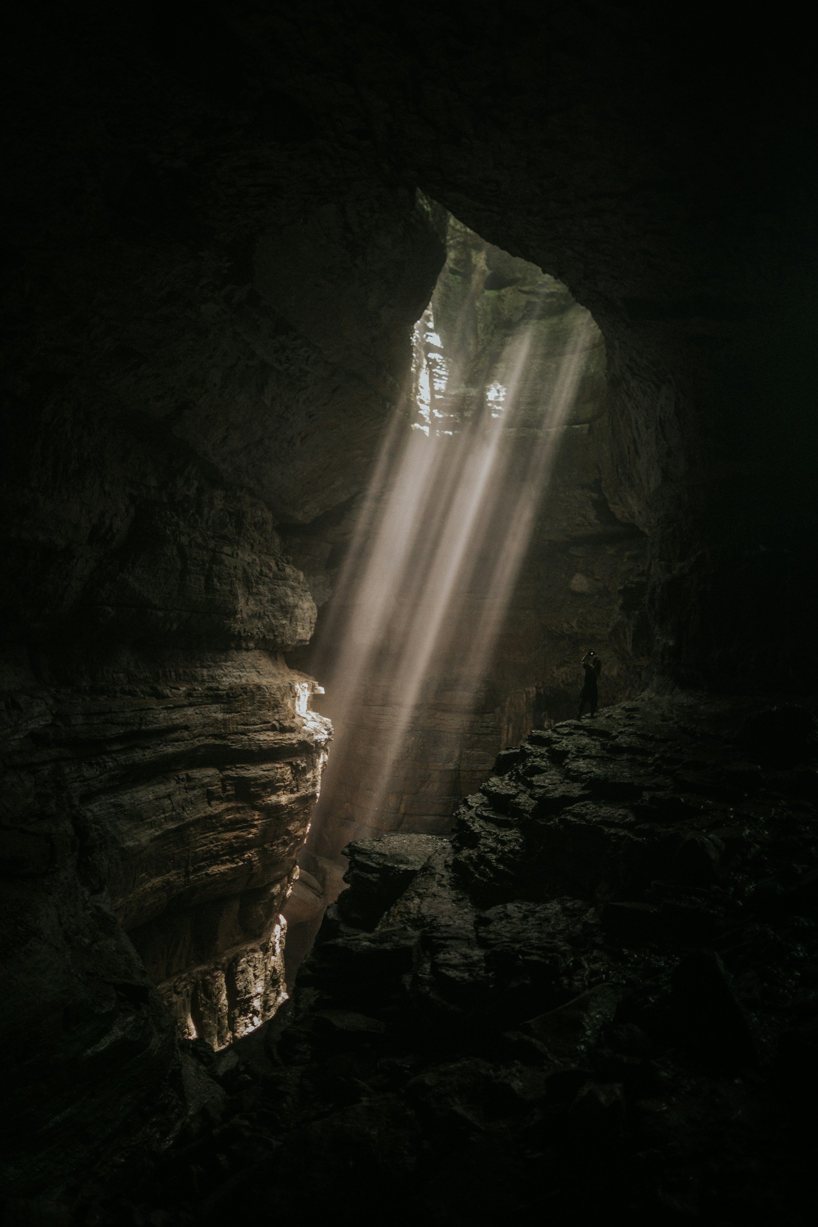 Sunlight streams into a dark cave through a hole in the ceiling, illuminating the rocky interior and a person standing on the cave floor.