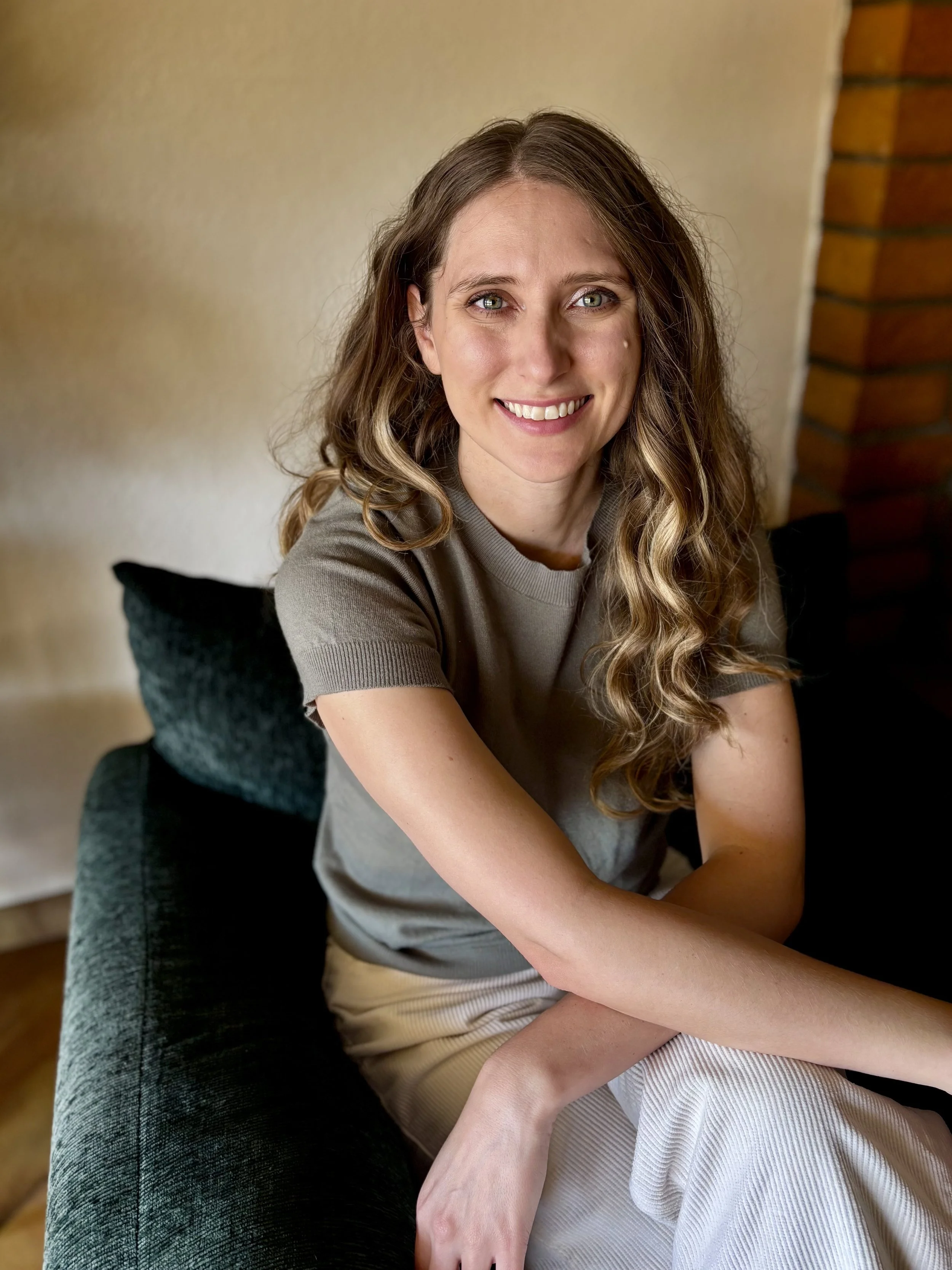 A woman with wavy light brown hair and blue eyes smiling while sitting on a dark green couch near a beige wall and a brick column.