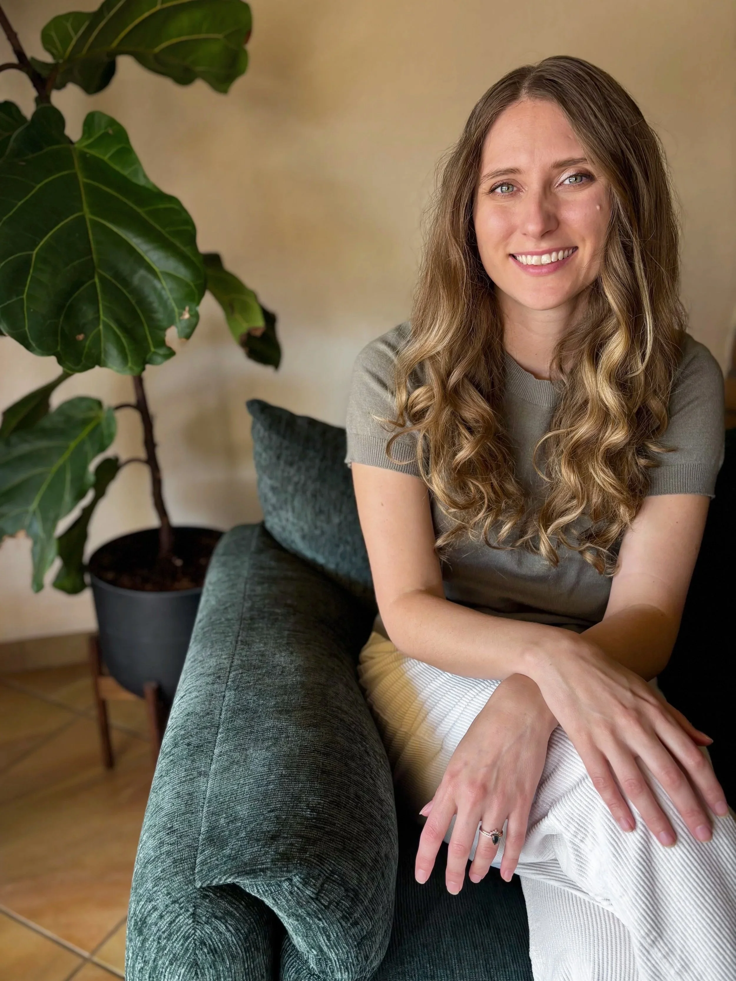 A woman with long, wavy, light brown hair smiling while sitting on a dark green sofa in a living room.