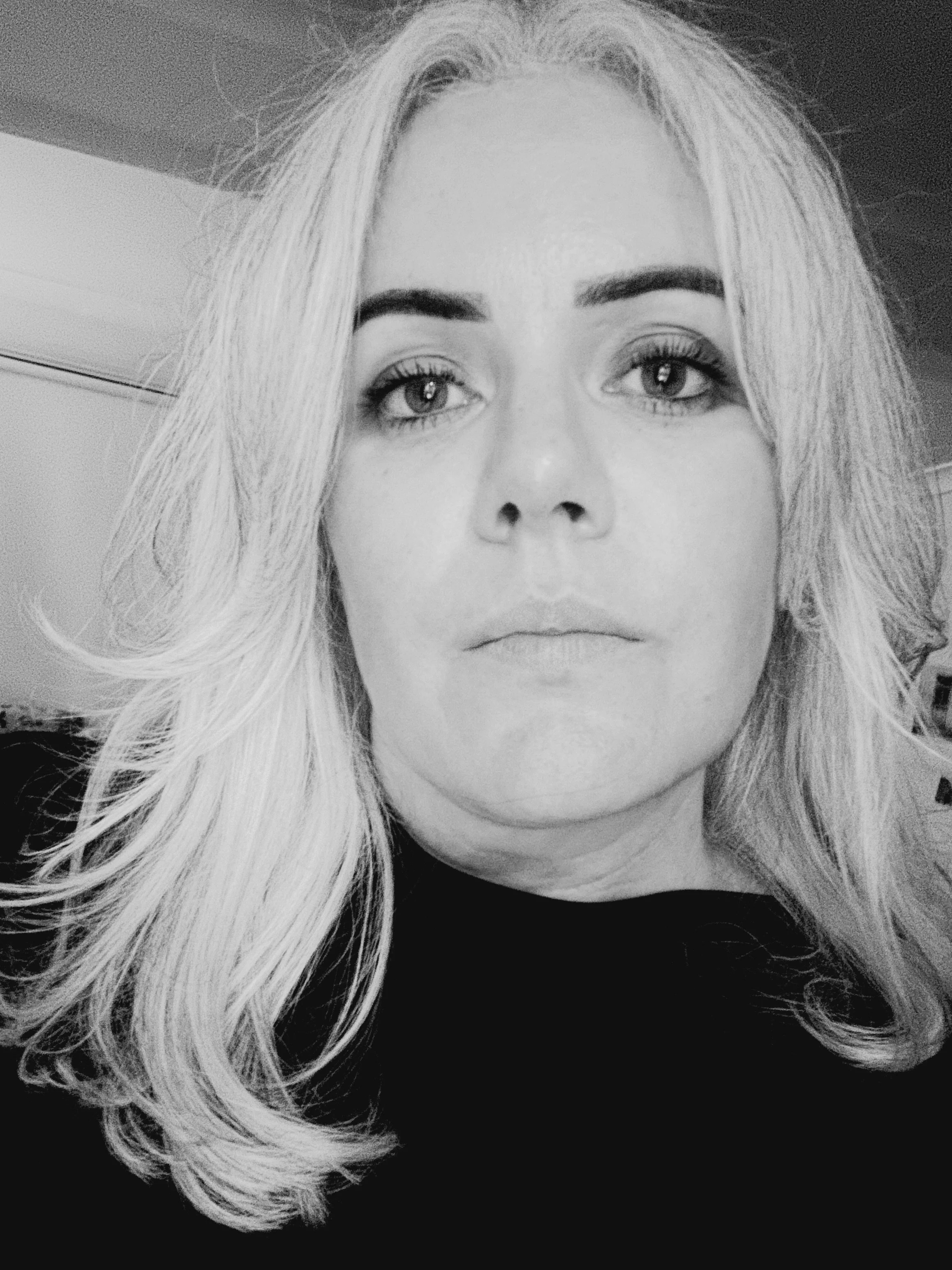 Close-up black and white photo of a woman with long, wavy silver hair, wearing a dark top, looking directly at the camera.