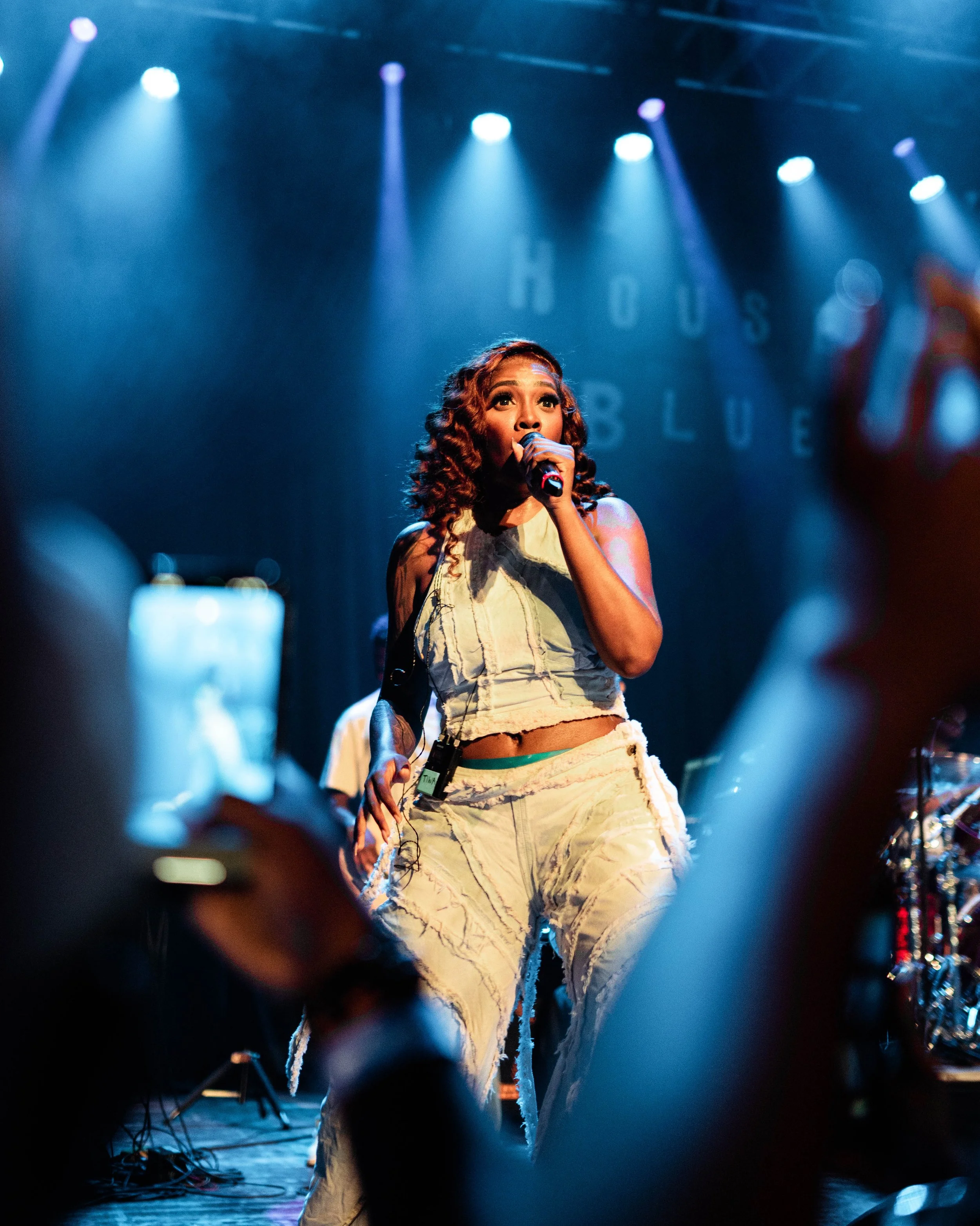 A female singer performs on stage, holding a microphone, with blue stage lights overhead and a backdrop that reads 'HOUSE' and 'BLUE'. She has curly hair and wears a sleeveless, light-colored outfit.
