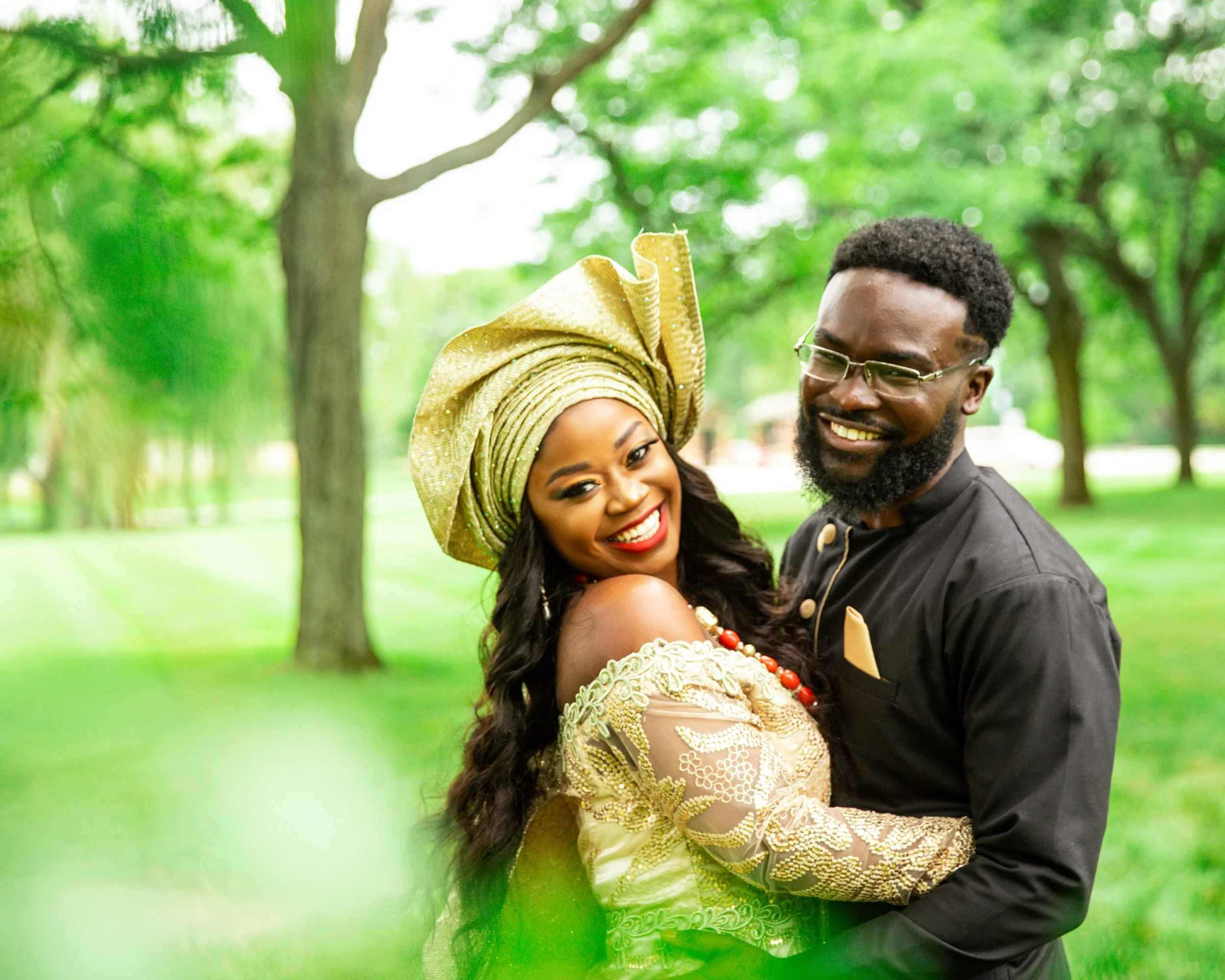 A smiling couple in traditional African attire, standing in a park with green trees in the background. The woman wears a gold and green outfit with a matching headwrap, and the man wears a black shirt with glasses.