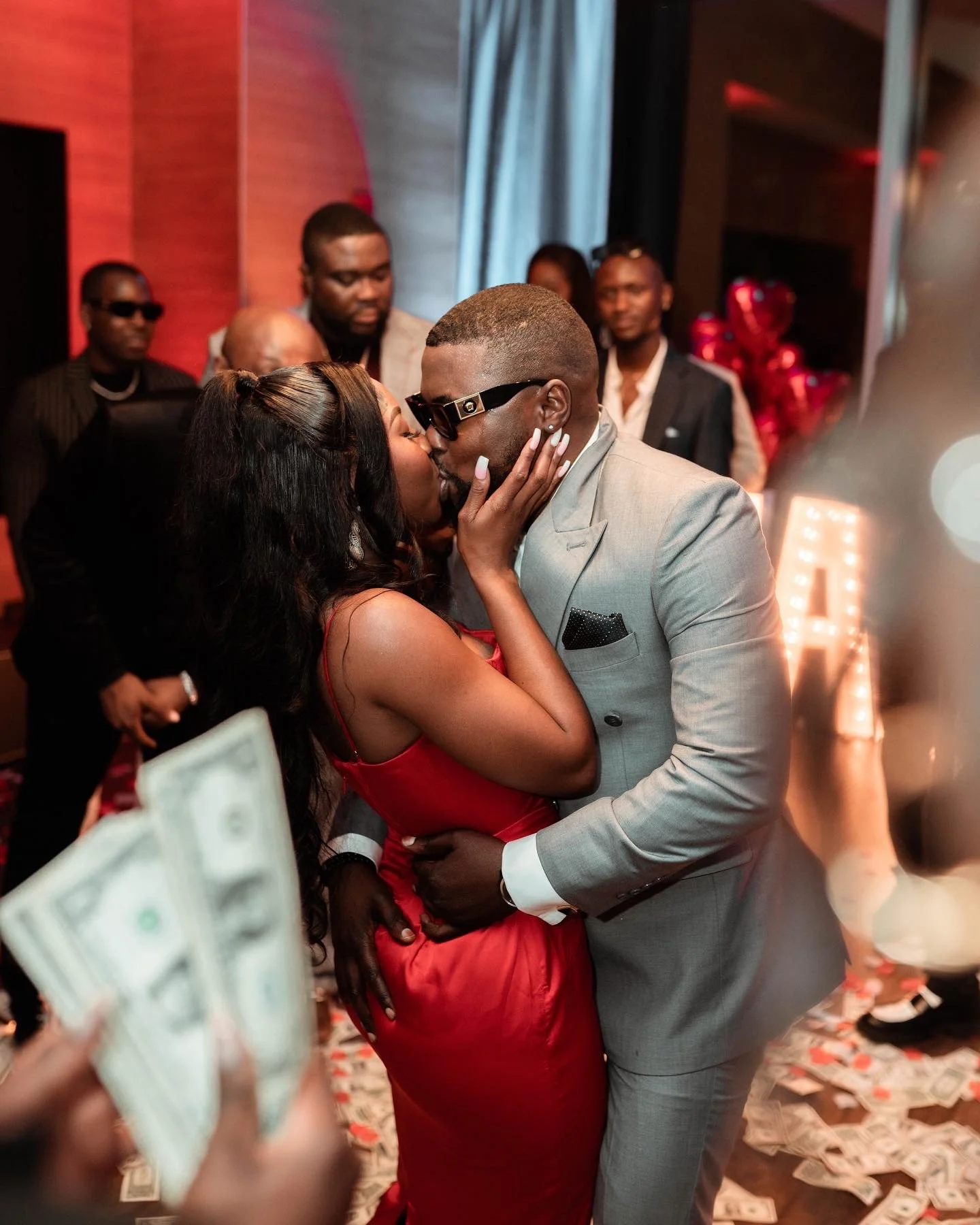 A couple is kissing at a celebration, surrounded by people, with dollar bills scattered on the floor and a decorative background. surprise proposal photoshoot