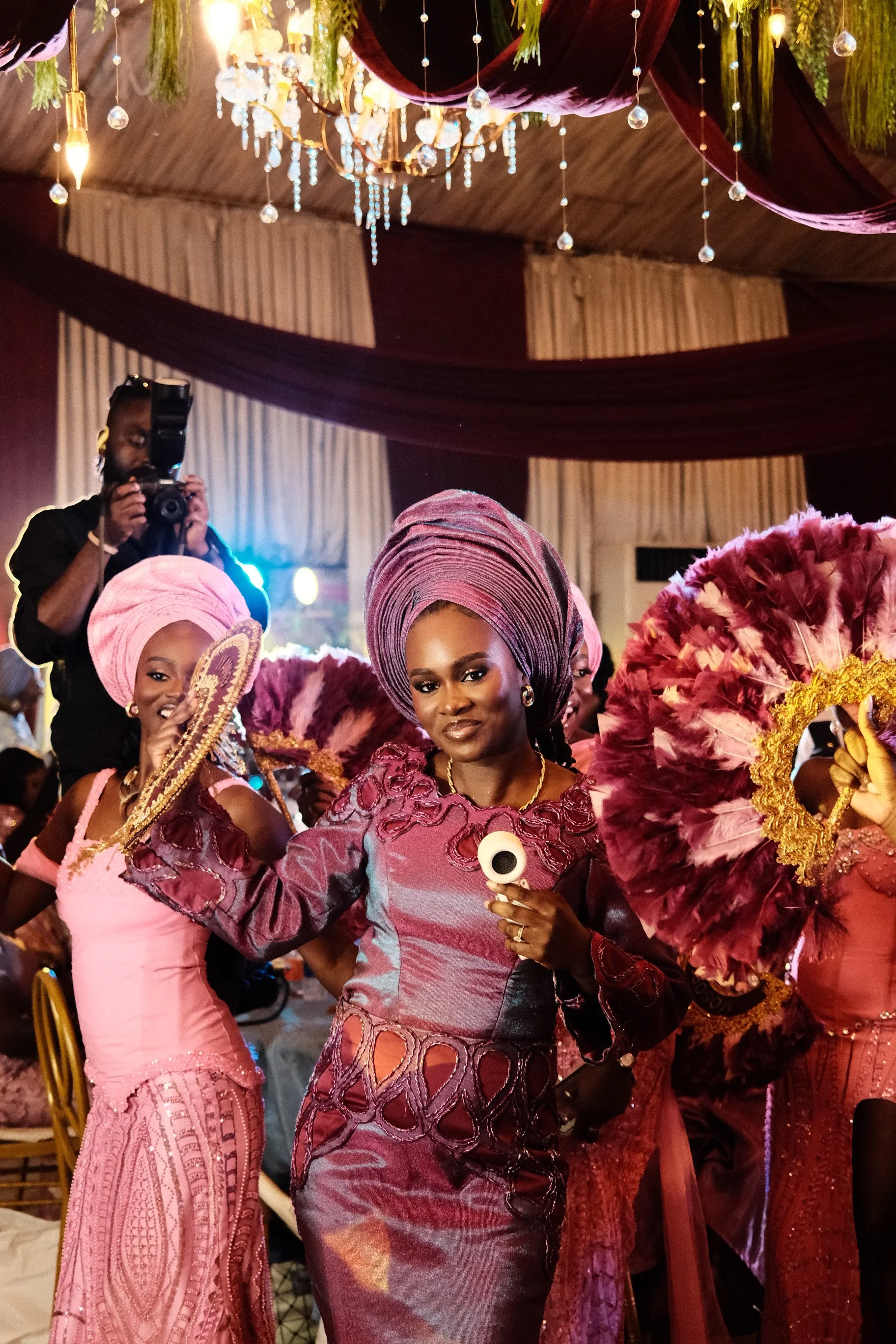 Women in traditional and ornate pink and purple attire at a celebration, with a chandelier and draped fabric in the background. wedding photography
