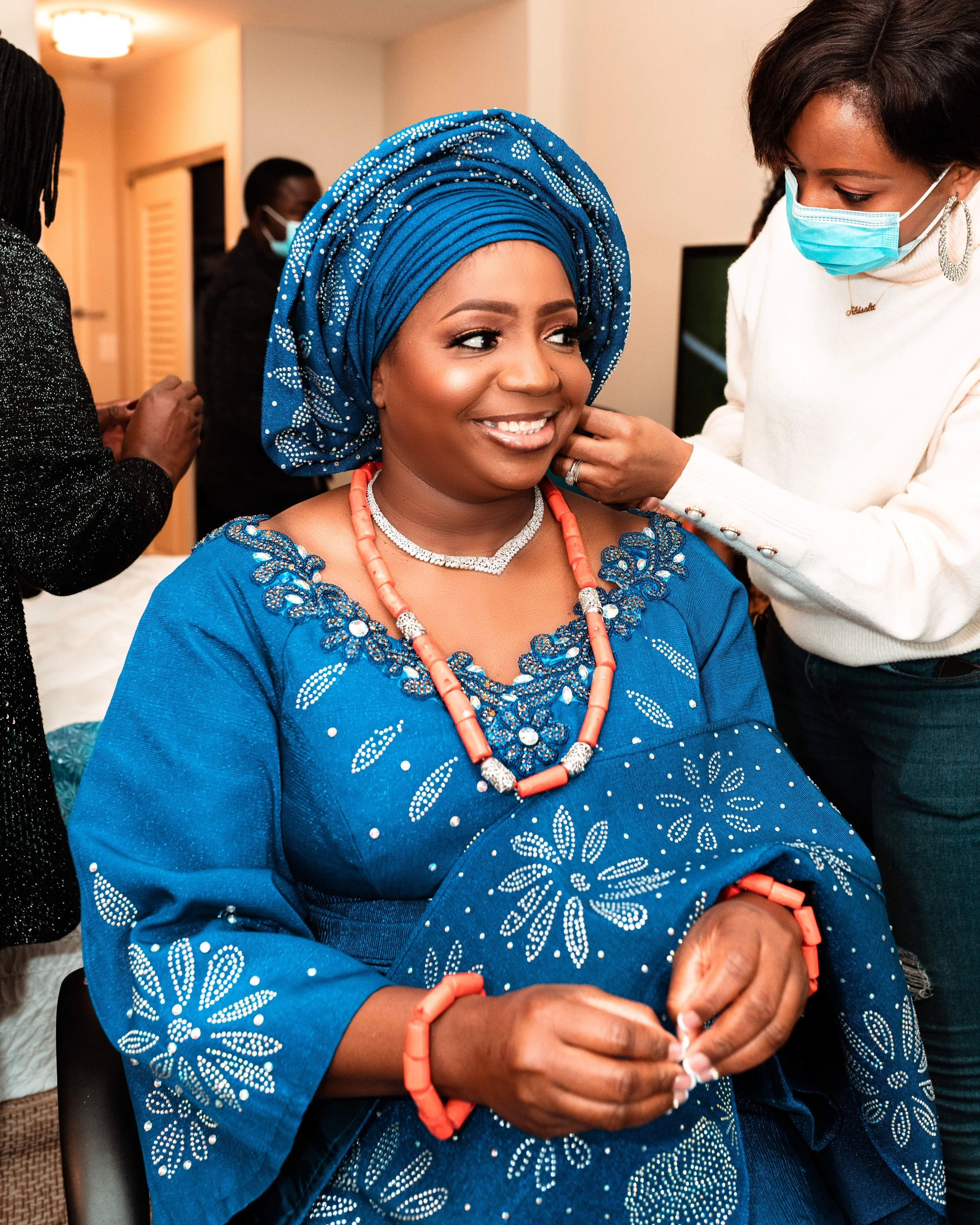 Woman dressed in traditional blue attire and headwrap, wearing coral and pearl jewelry, smiling while getting her earrings adjusted by a woman in a white sweater and face mask. Other people in the background.