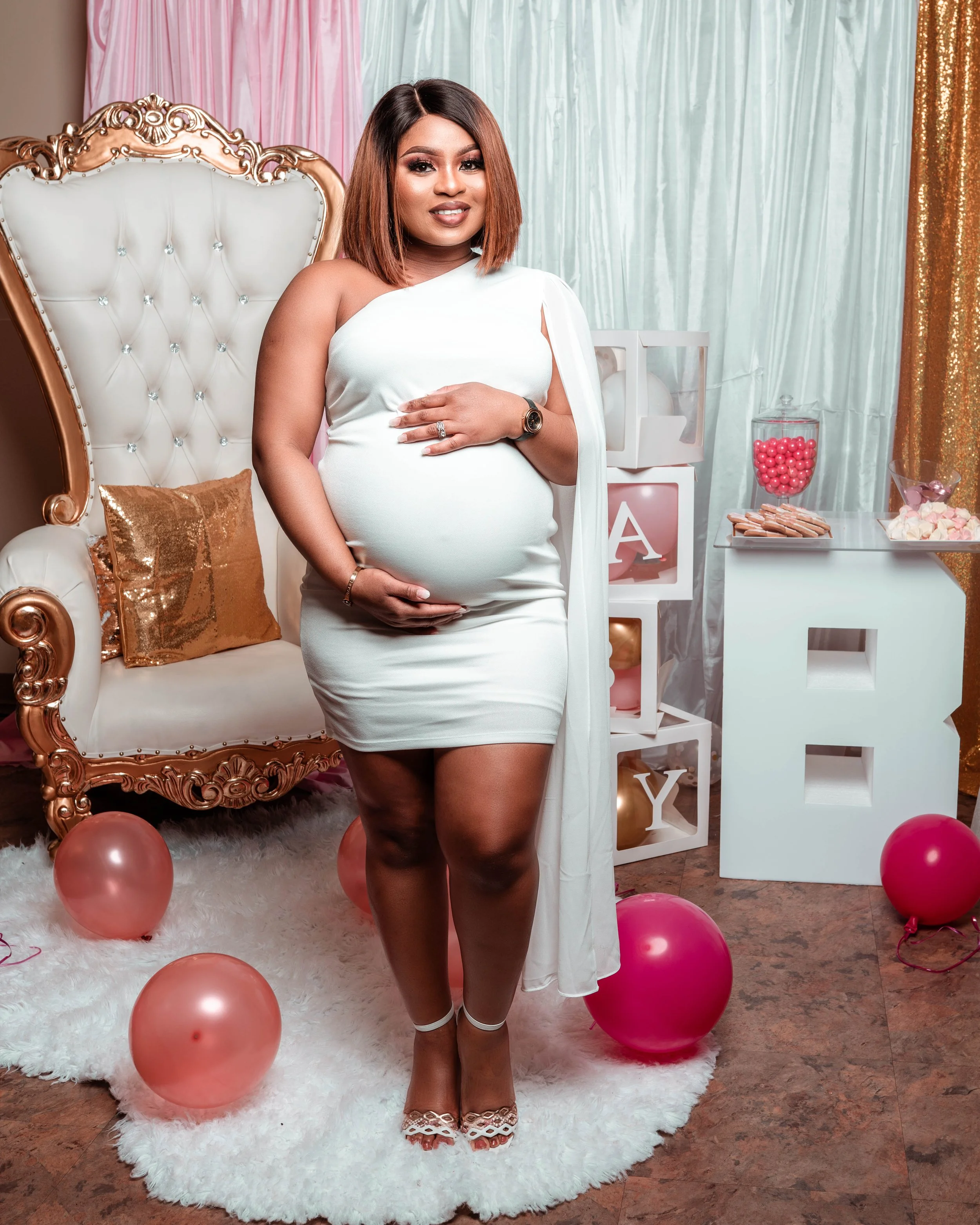 A pregnant woman in a white dress standing in a decorated room with pink and gold balloons, a plush white rug, a gold and white ornate chair, and a backdrop with pastel-colored curtains, celebrating a gender reveal or baby shower.