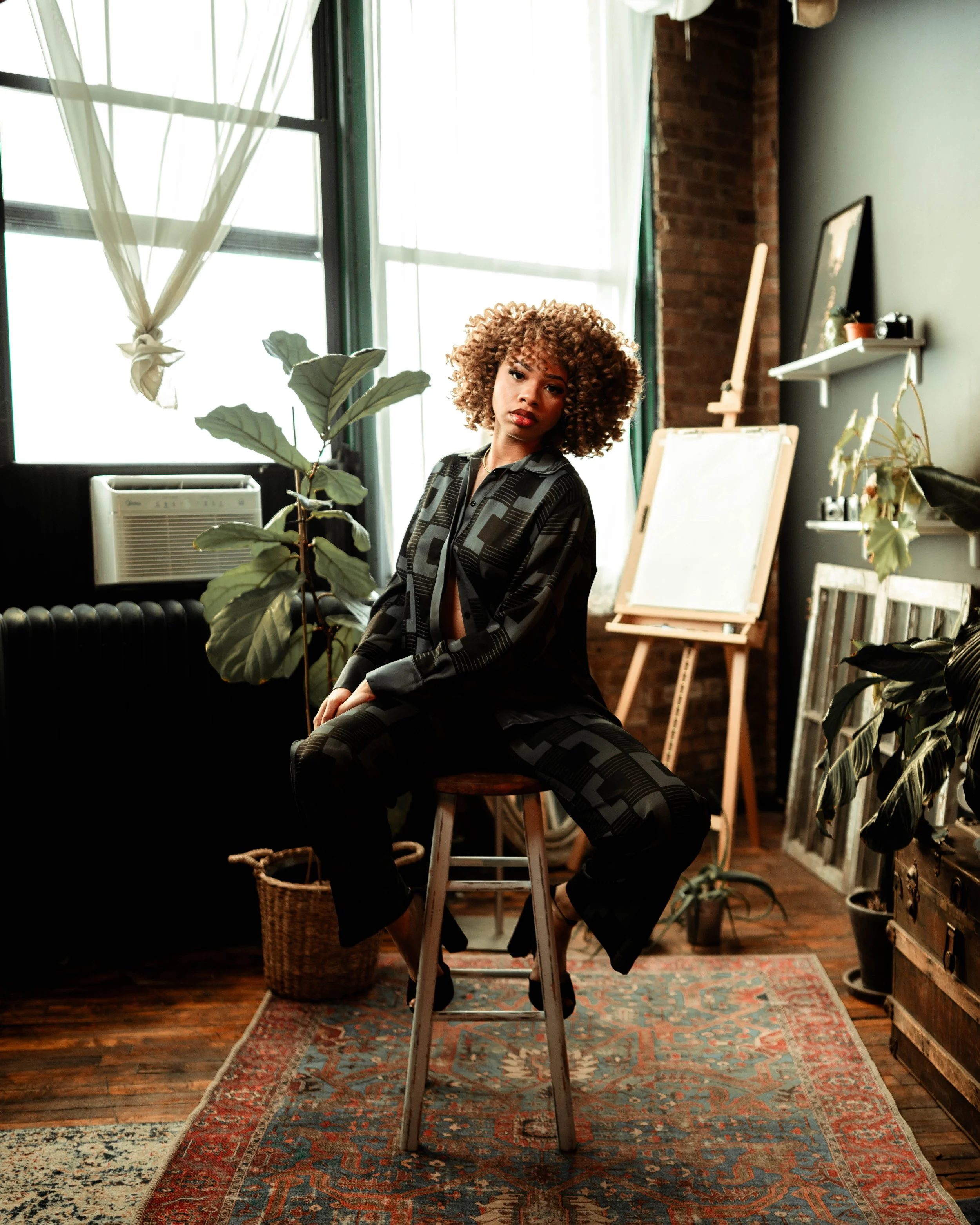 A woman with curly hair seated on a wooden stool in an art studio or gallery with natural light, plants, and art supplies in the background.