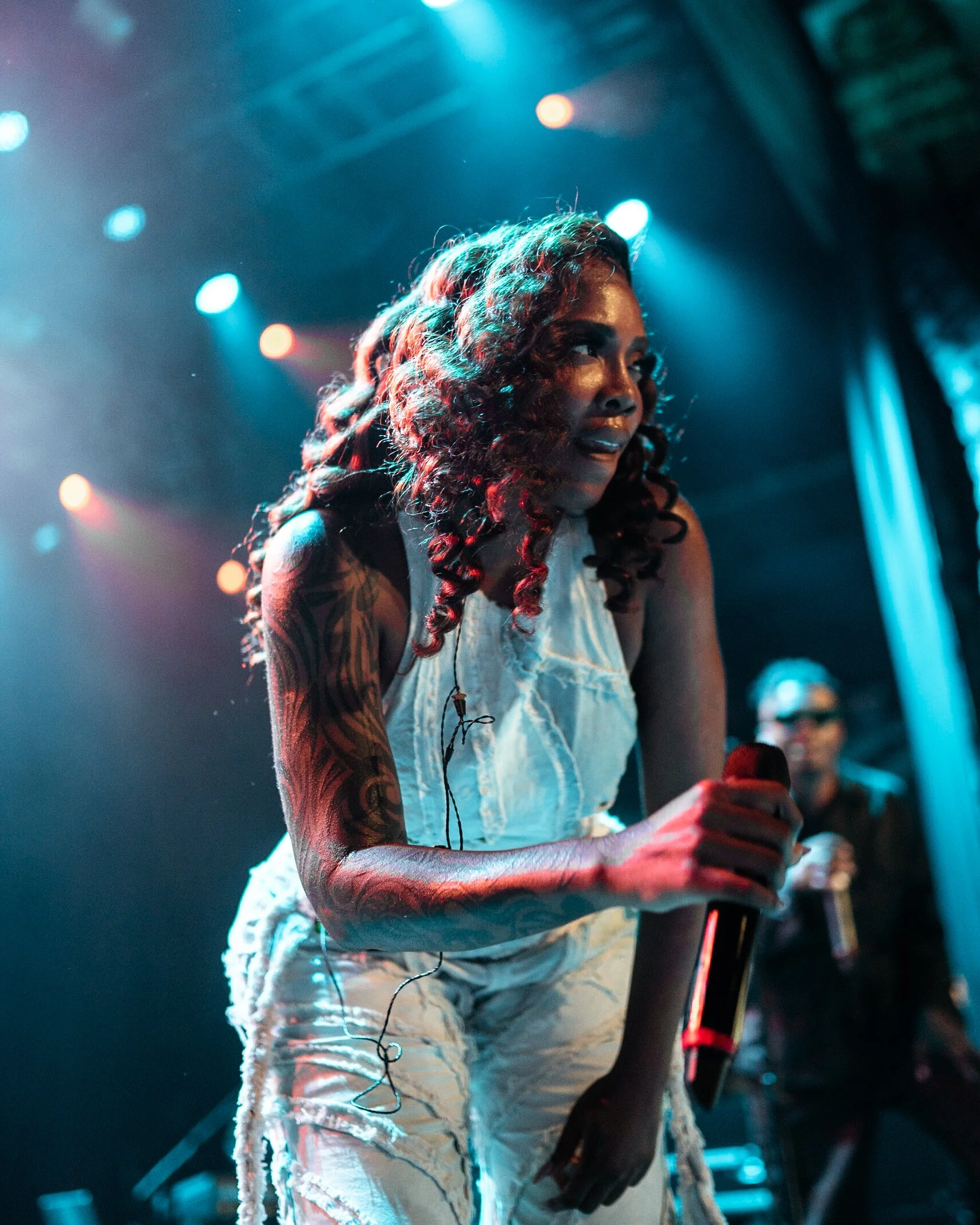 A woman with curly hair and tattoos holding a microphone on stage, illuminated by colorful concert lighting.