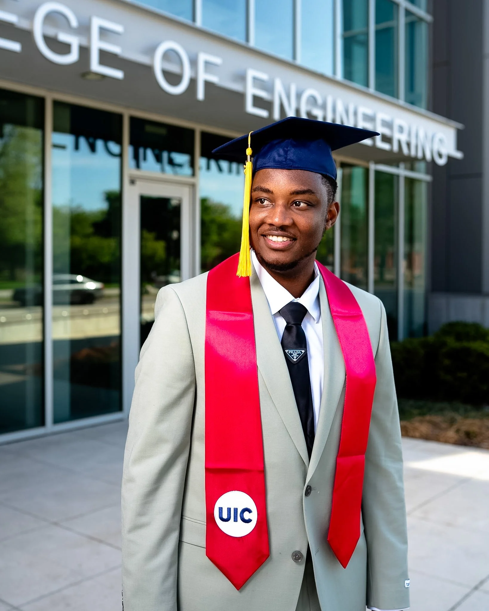 Young man in a beige suit and graduation cap with a yellow tassel, wearing a pink honor cord and UIC badge, standing outside a building with 'College of Engineering' sign. graduation photoshoot