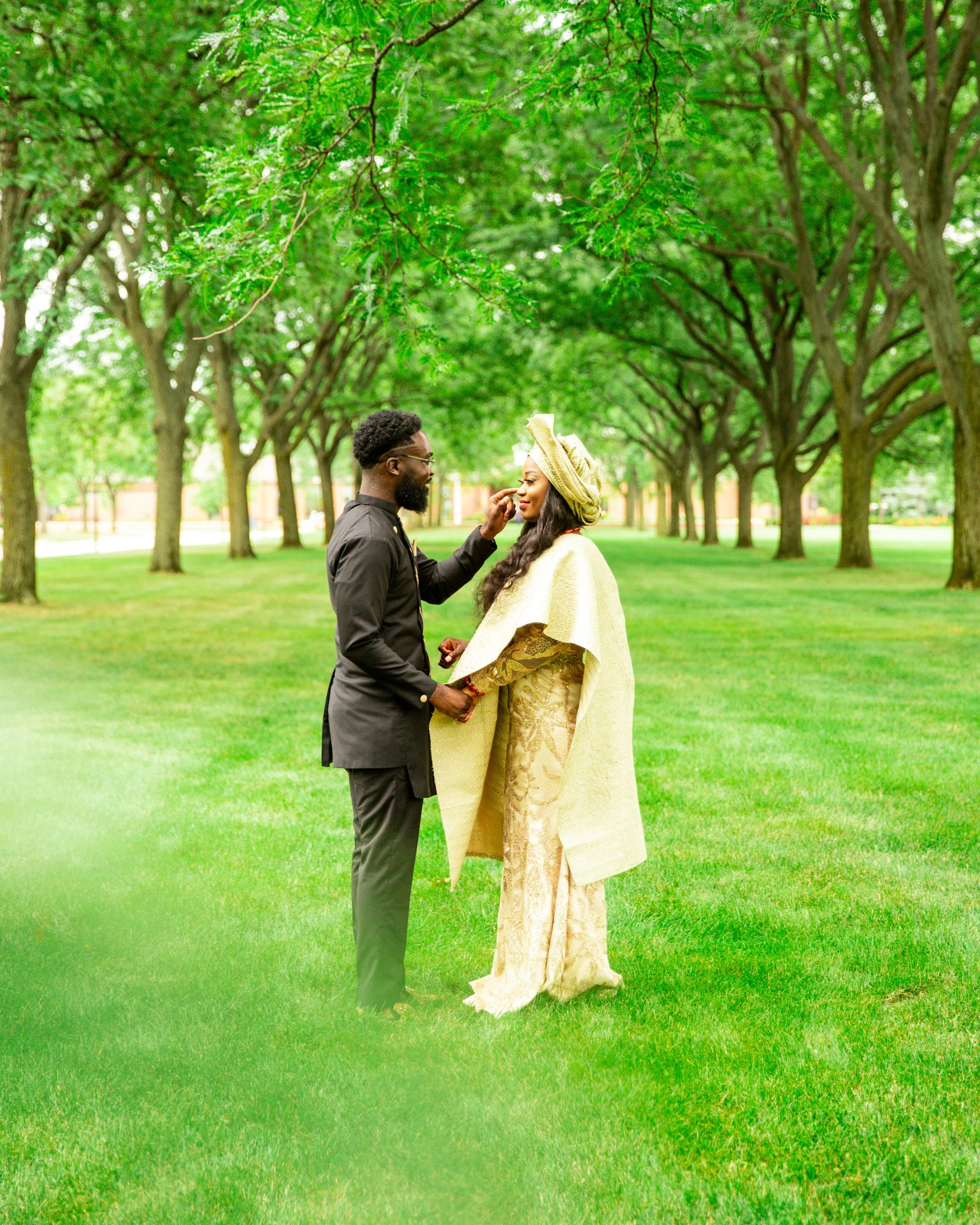 A couple dressed in elegant attire standing in a lush green park with tall trees. The man wearing a black outfit and glasses is touching the woman's face, who is wearing a gold traditional dress with a matching hat. They are holding hands and sharing