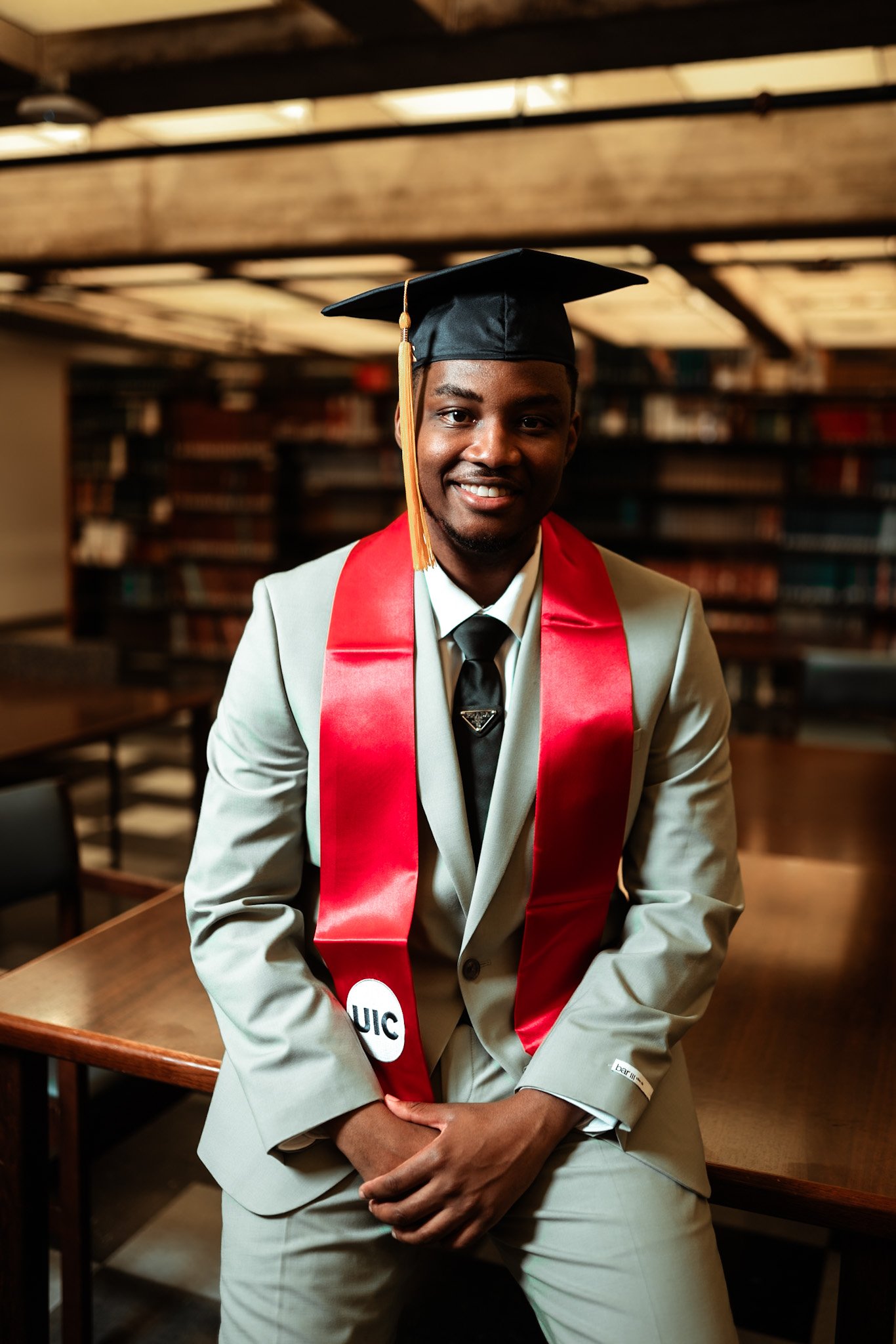 A young man in a beige suit with a black tie, wearing a graduation cap and red stole, is smiling and sitting in a library or study room. Graduation photoshoot