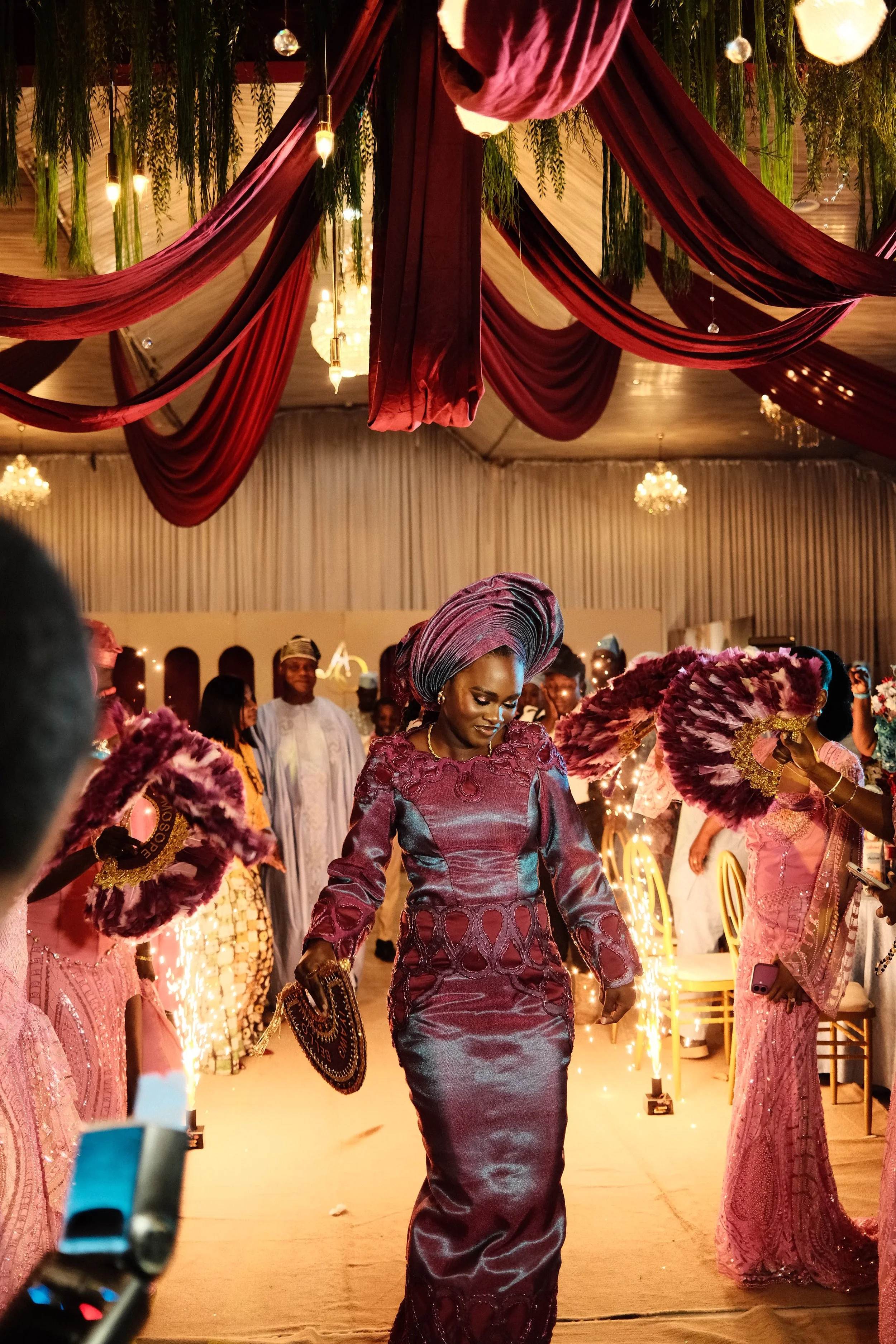Women in traditional Nigerian attire at a celebration, with a woman in the center wearing a purple agbada and gele headwrap, surrounded by others holding feather fans, in a decorated event hall with curtains, hanging lights, and chandeliers.