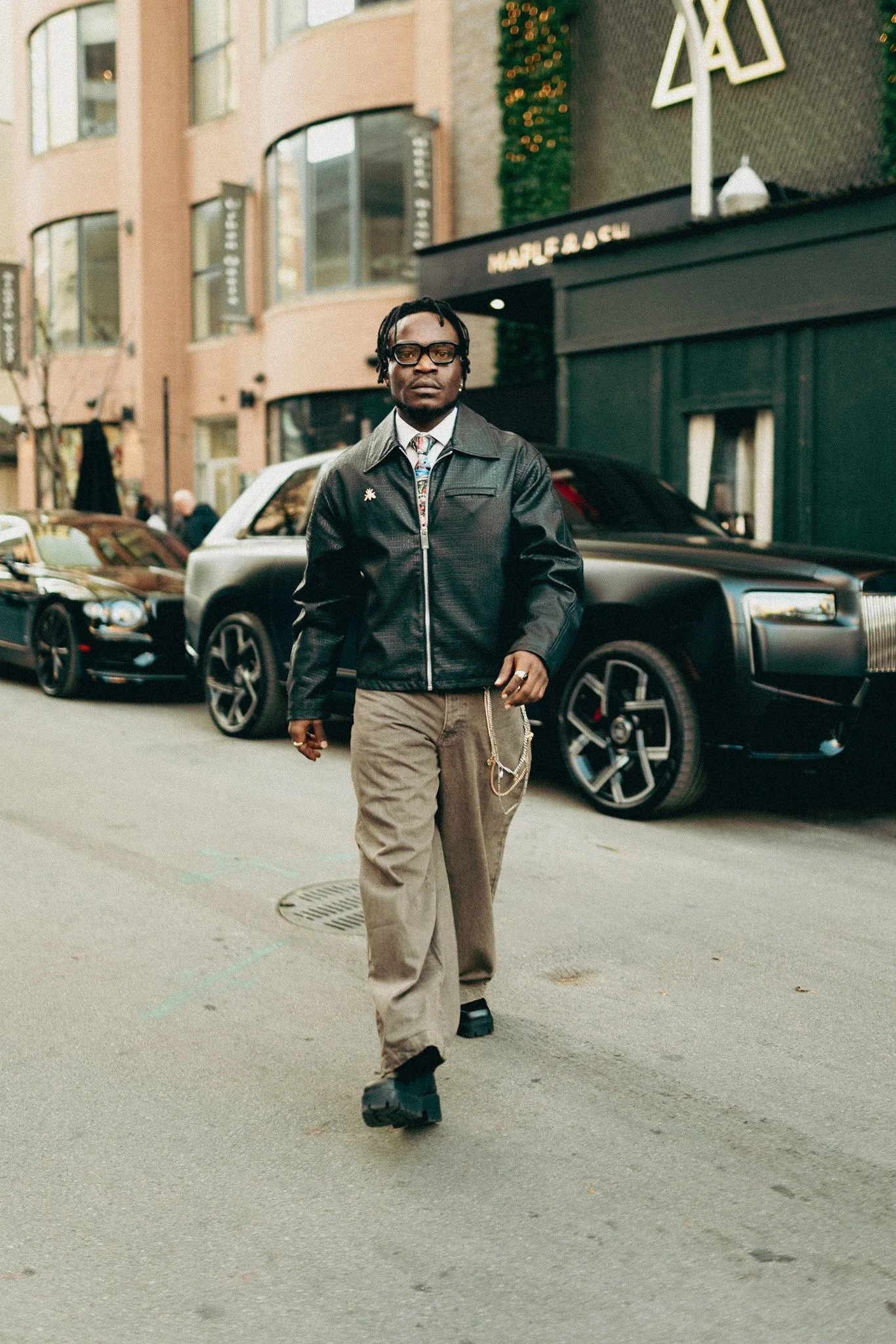 A man with glasses, dreadlocks, and a dark jacket walking on a city street with luxury cars parked behind him.