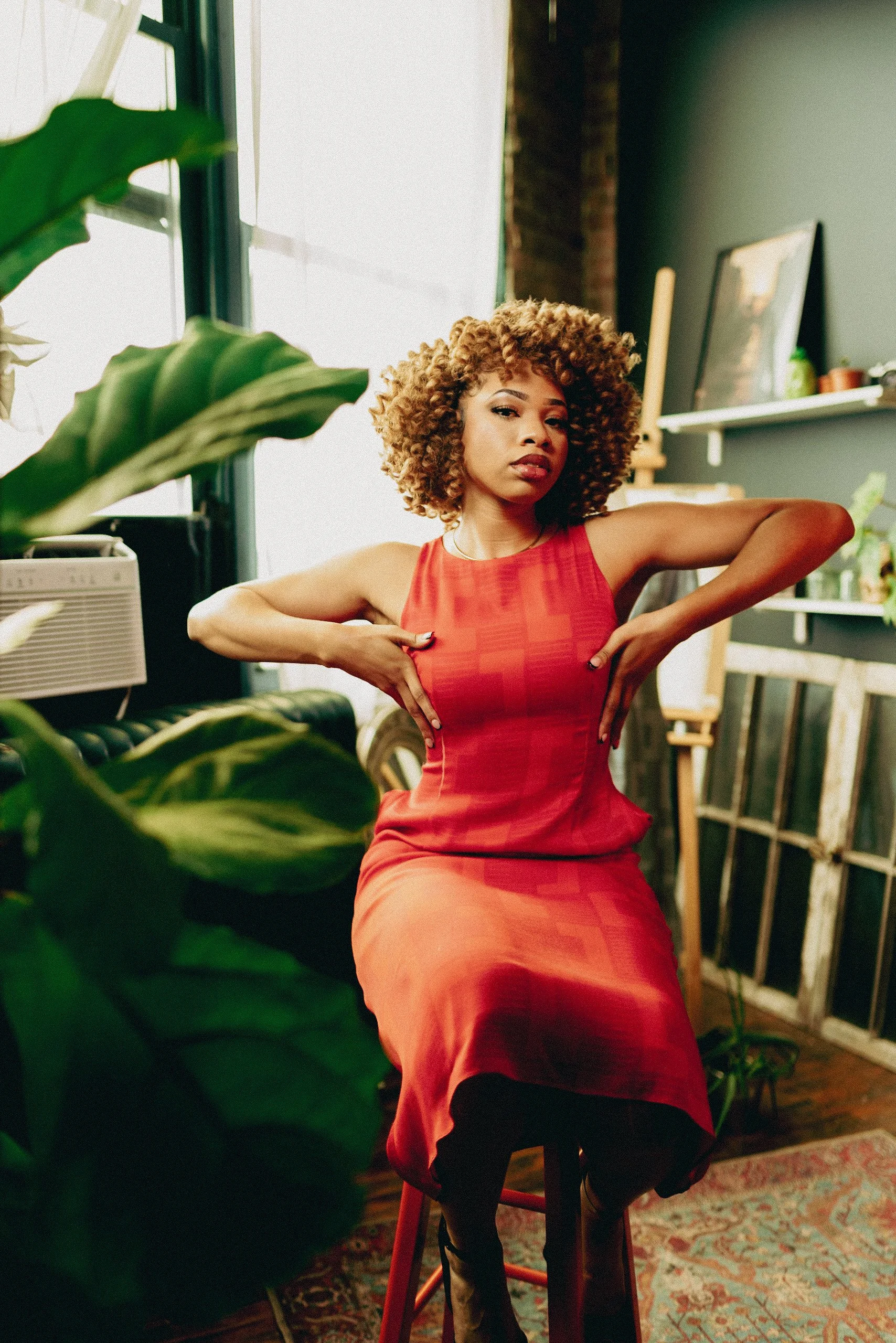 A woman with curly hair wearing a red dress sitting on a stool in an artist's studio or creative space, surrounded by plants, art supplies, and framed artwork.