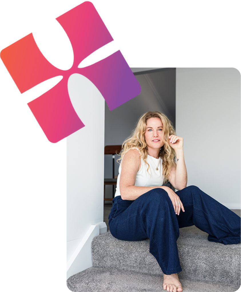 A woman with long blonde wavy hair sitting on carpeted stairs in a modern home, wearing a white sleeveless top and dark blue wide-leg jeans, with a neutral expression.