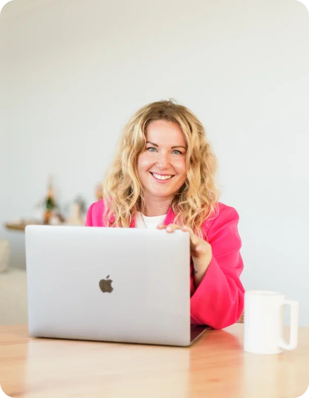 A smiling woman with blonde curly hair wearing a bright pink blazer, sitting at a wooden table with a silver Apple MacBook and a white mug in front of her, in a well-lit room.