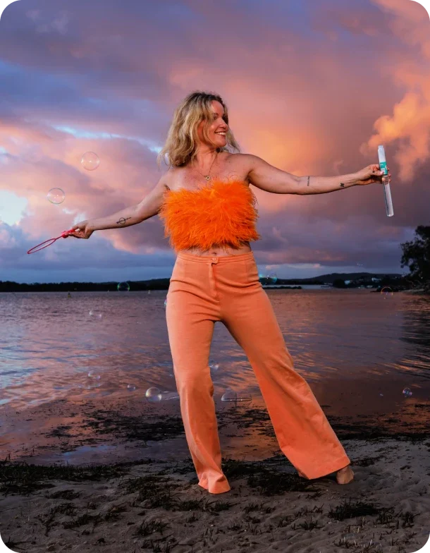 A woman wearing an orange feather top and high-waisted orange pants standing on a beach at sunset, blowing bubbles.
