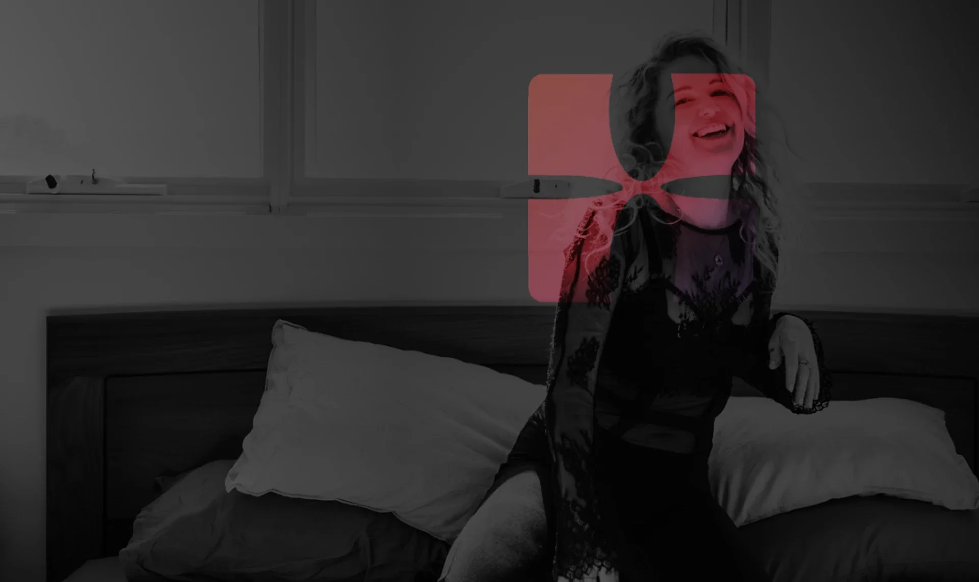 A woman with curly hair laughing while sitting on a bed in a bedroom, with window behind her, monochrome photo with a red overlay.