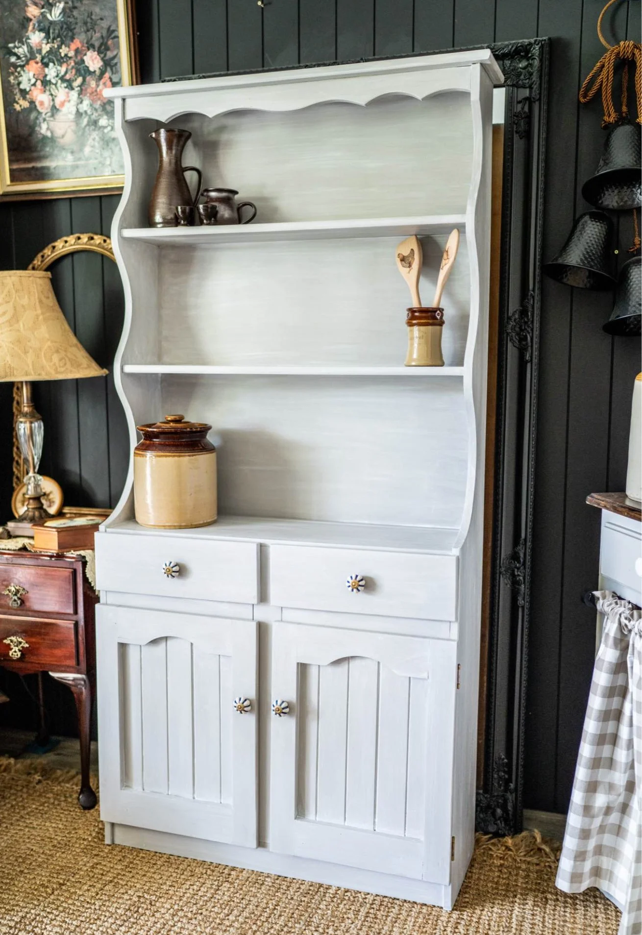 A white vintage-style wooden cabinet with open shelves and two drawers, decorated with ceramic and wood kitchenware, placed in a room with dark paneled walls, a gold-framed floral painting, a traditional table lamp, and a nearby small wooden table.