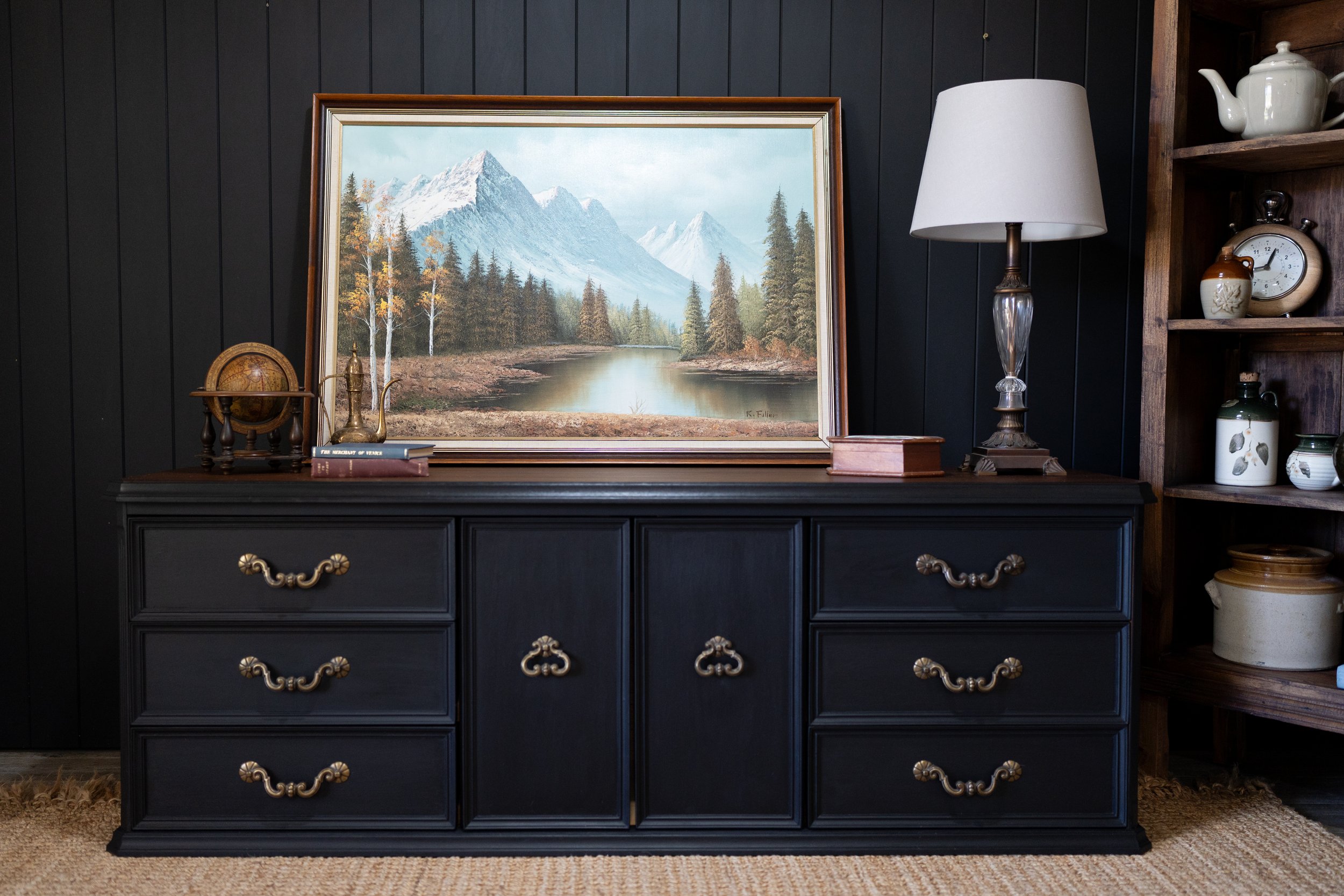 A black wooden dresser with gold-colored hardware, topped by a framed landscape painting of mountains, trees, and a river, a small globe, a brass teapot, and two books. To the right, a white table lamp with a glass base is next to a dark wood shelf with decorative pottery and a vintage clock. The background features a black wooden wall and a beige carpet.