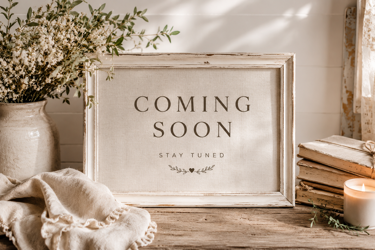 A decorative sign with the words 'Coming Soon, Stay Tuned' surrounded by a rustic setting including a vase of white flowers, a stack of old books, a lit candle, and a beige cloth on a wooden surface.