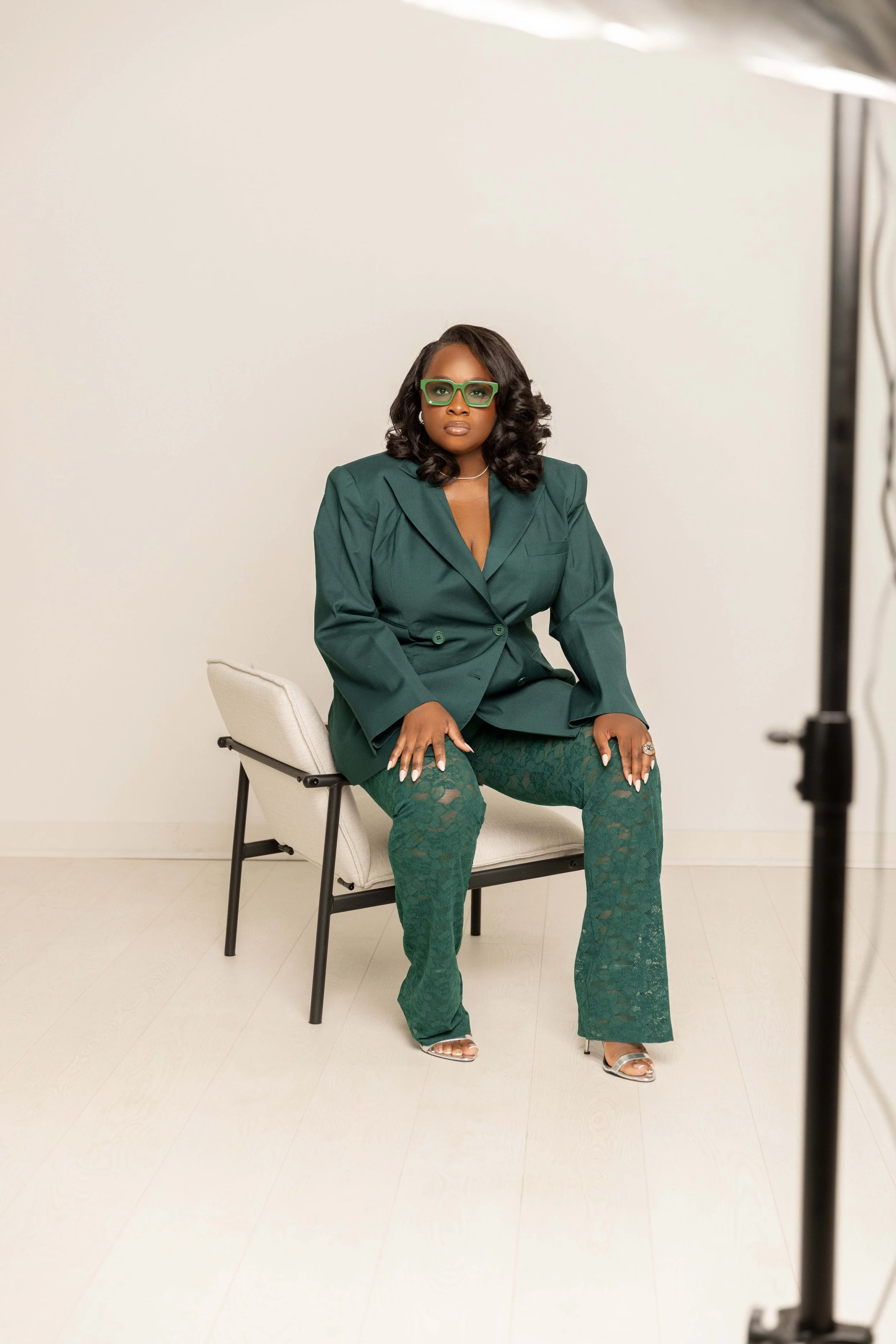 A woman sitting on a chair in a photo studio, wearing a green suit and lace pants, with green glasses and silver heels, looking serious and confident.