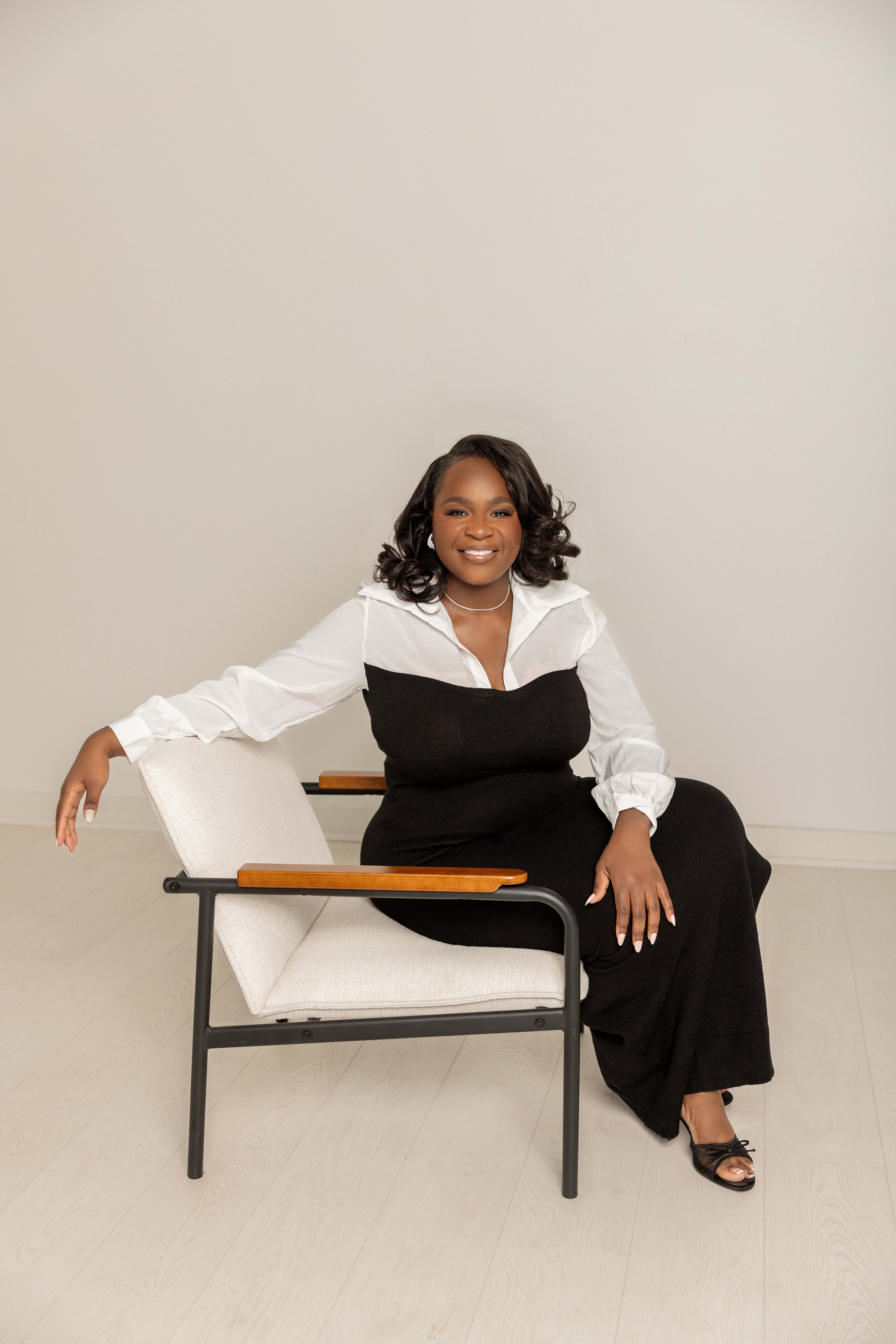 A woman with dark hair styled in loose waves is sitting on a white chair with black metal legs and wooden armrests, smiling at the camera. She is wearing a white blouse with a black dress or sweater over it, and black high-heeled sandals. She is seated in front of a plain beige wall in a brightly lit room.