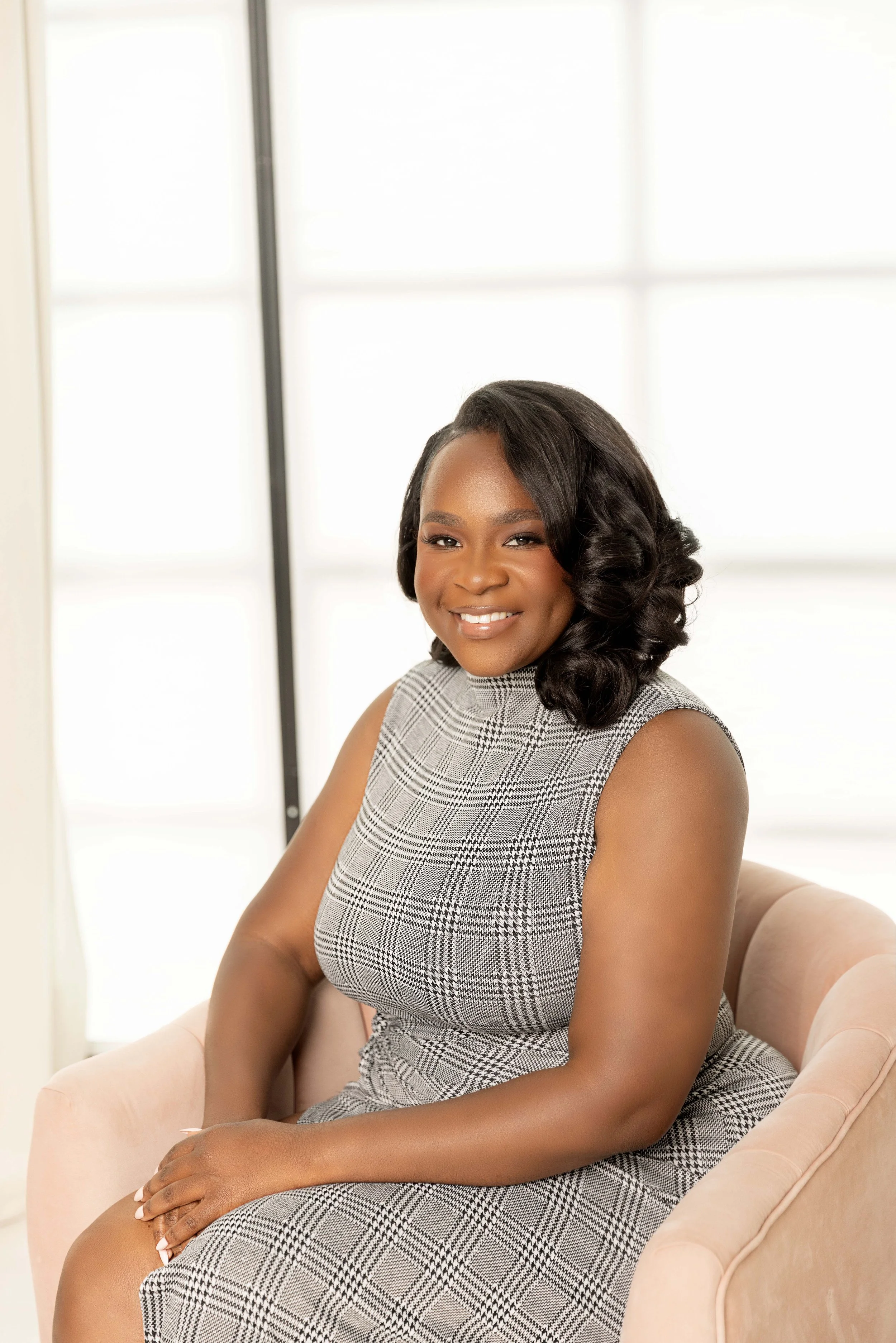 Smiling woman sitting on a pink chair in front of a bright window, wearing a sleeveless gray checkered dress.