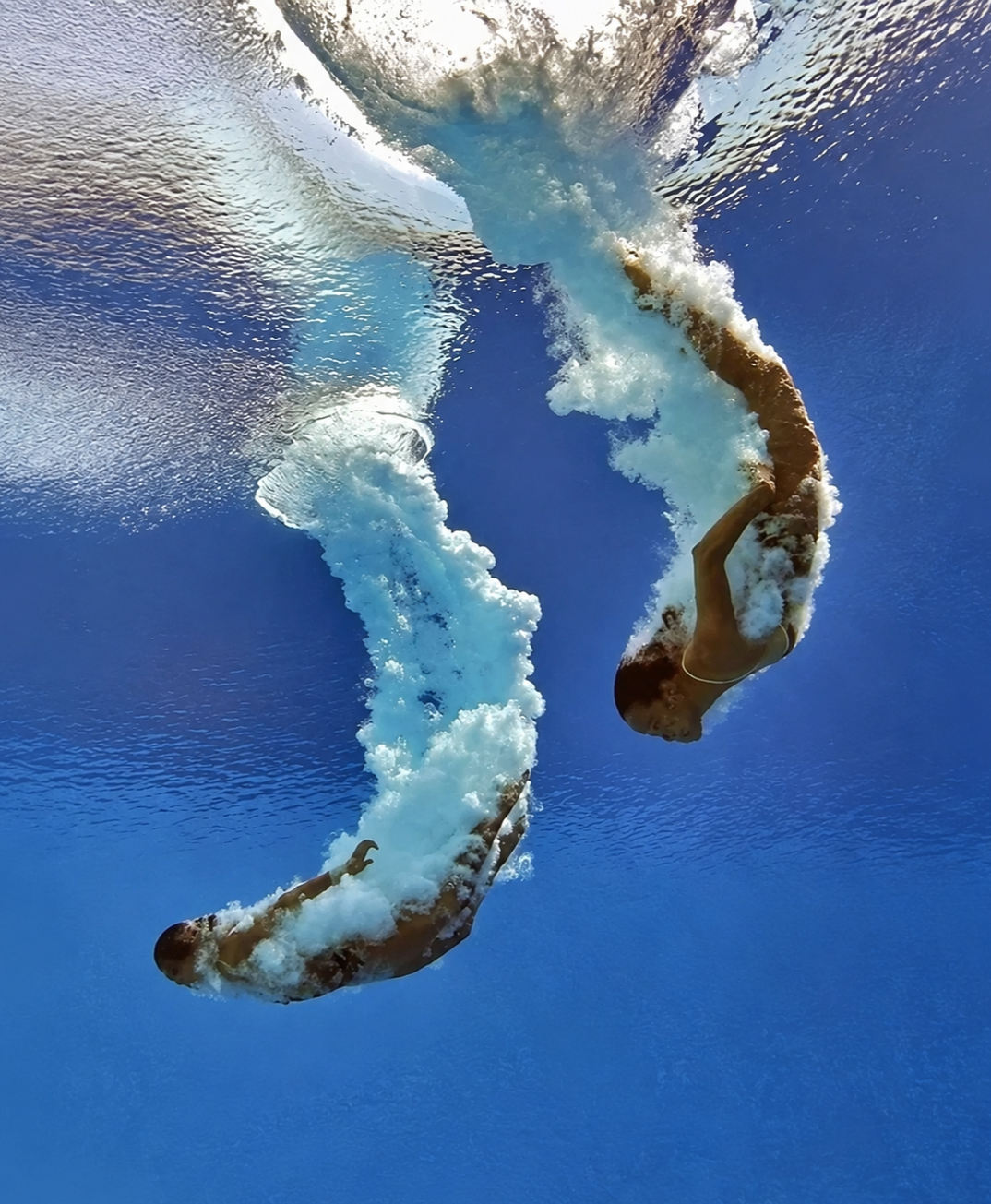 Two synchronized swimmers performing underwater in a pool, creating bubbles and ripples in the water.