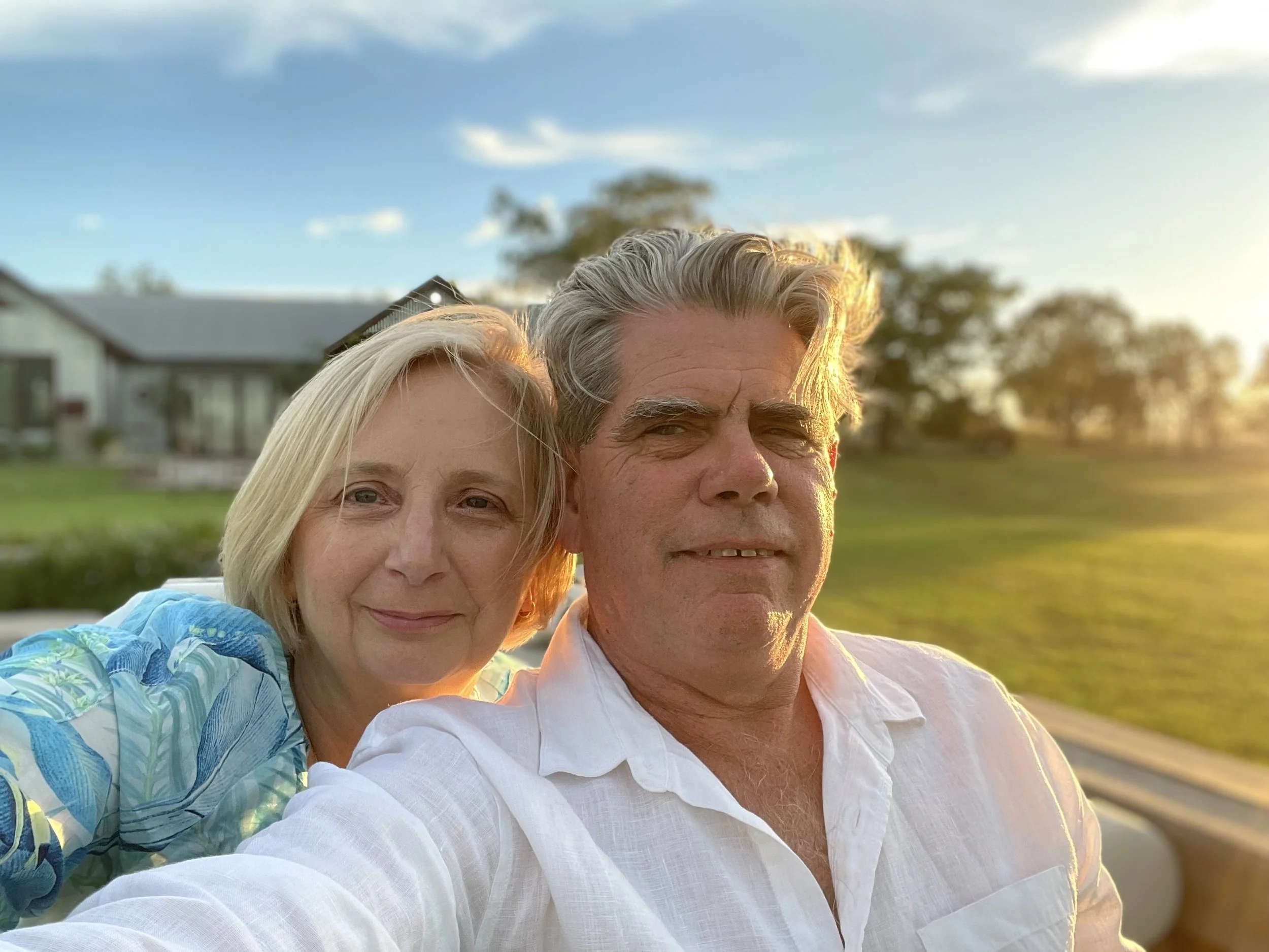 A middle-aged man and woman taking a selfie outdoors during sunset, with a house, trees, and a grassy yard in the background.