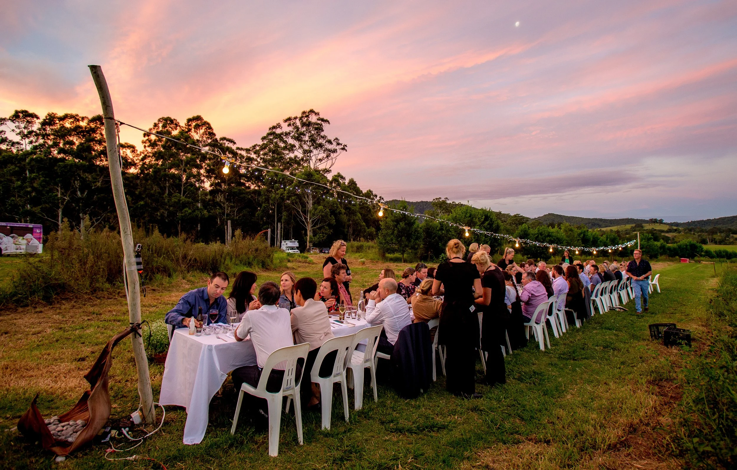 Dining outdoor gathering with many people seated at long tables under string lights at sunset in a rural landscape.