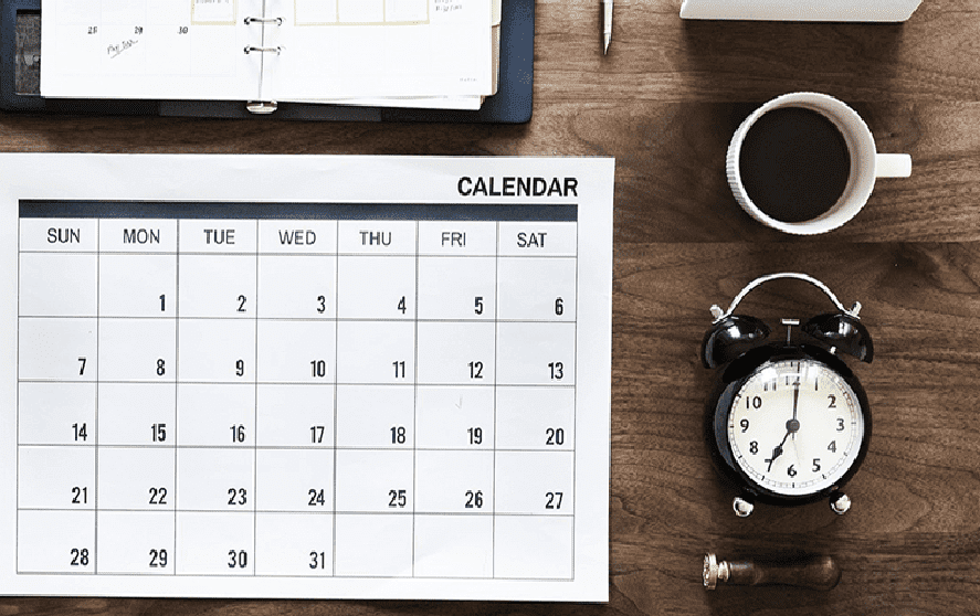 A desk with a calendar, a cup of coffee, a small black and white alarm clock, a seal stamp, and a planner.