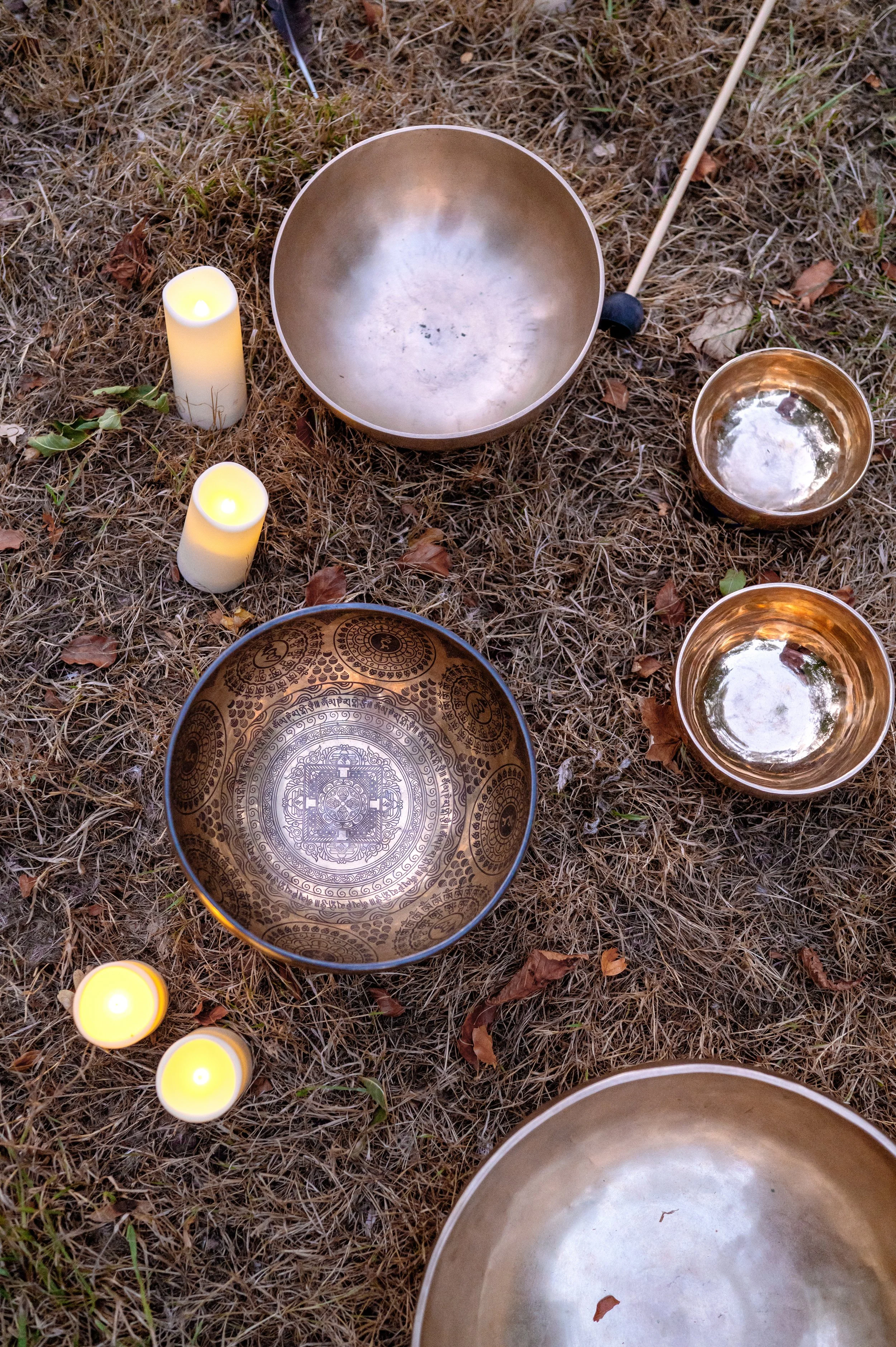 Three lit candles, two singing bowls, and one prayer bowl placed on dry grass outdoors.