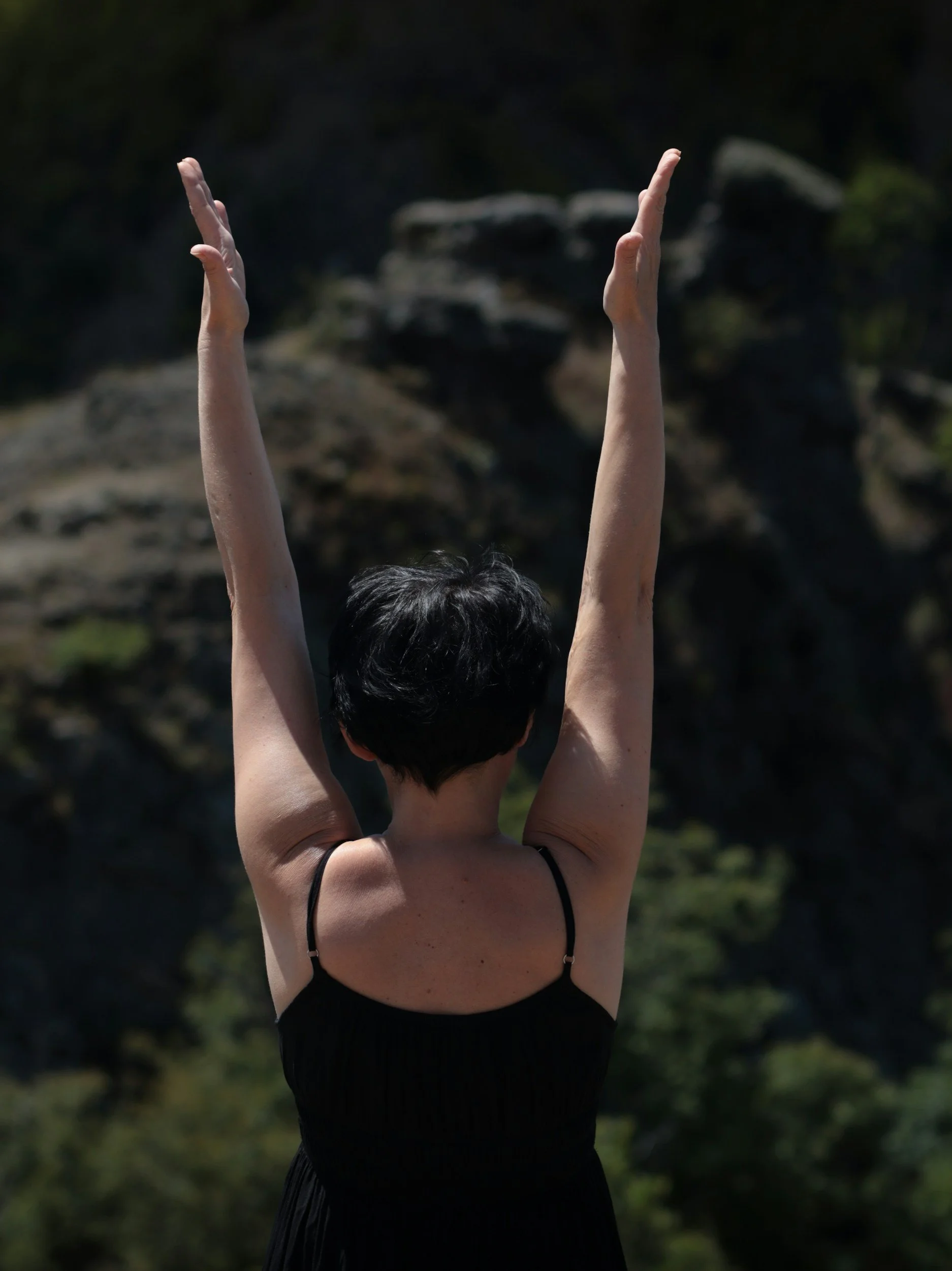 A person with short dark hair and bare arms, seen from the back, wearing a black sleeveless top and black skirt, raising their arms upward with hands facing outward, outdoors with dark rocks and greenery in the background.