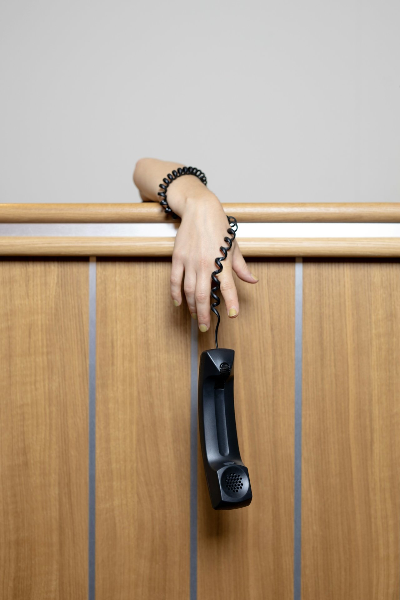 A hand rests on a wooden surface with a black corded telephone hanging from it. The person is waiting to talk to someone or be called or suggests contact us.