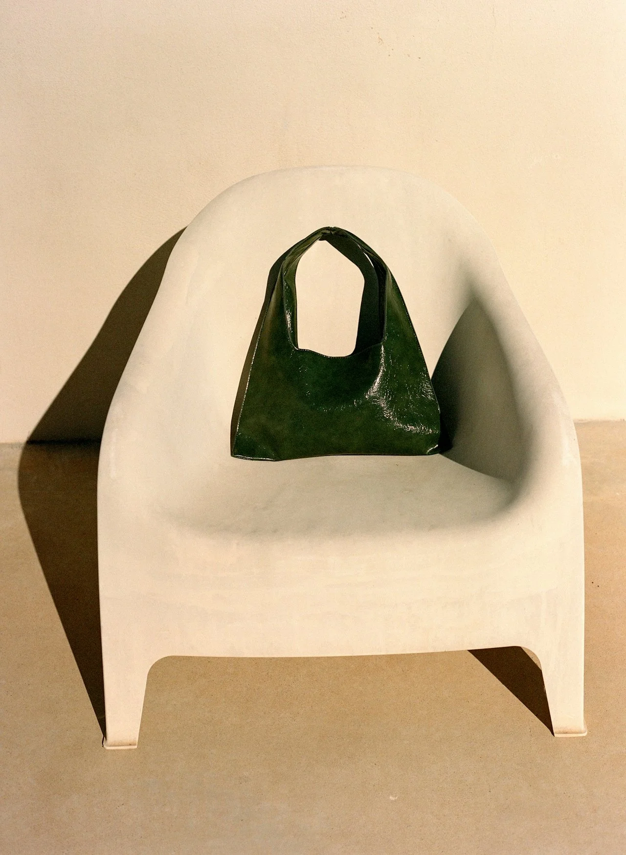 A small, shiny green shopping bag placed on a mid-century modern chair with a curved backrest against a beige wall.
