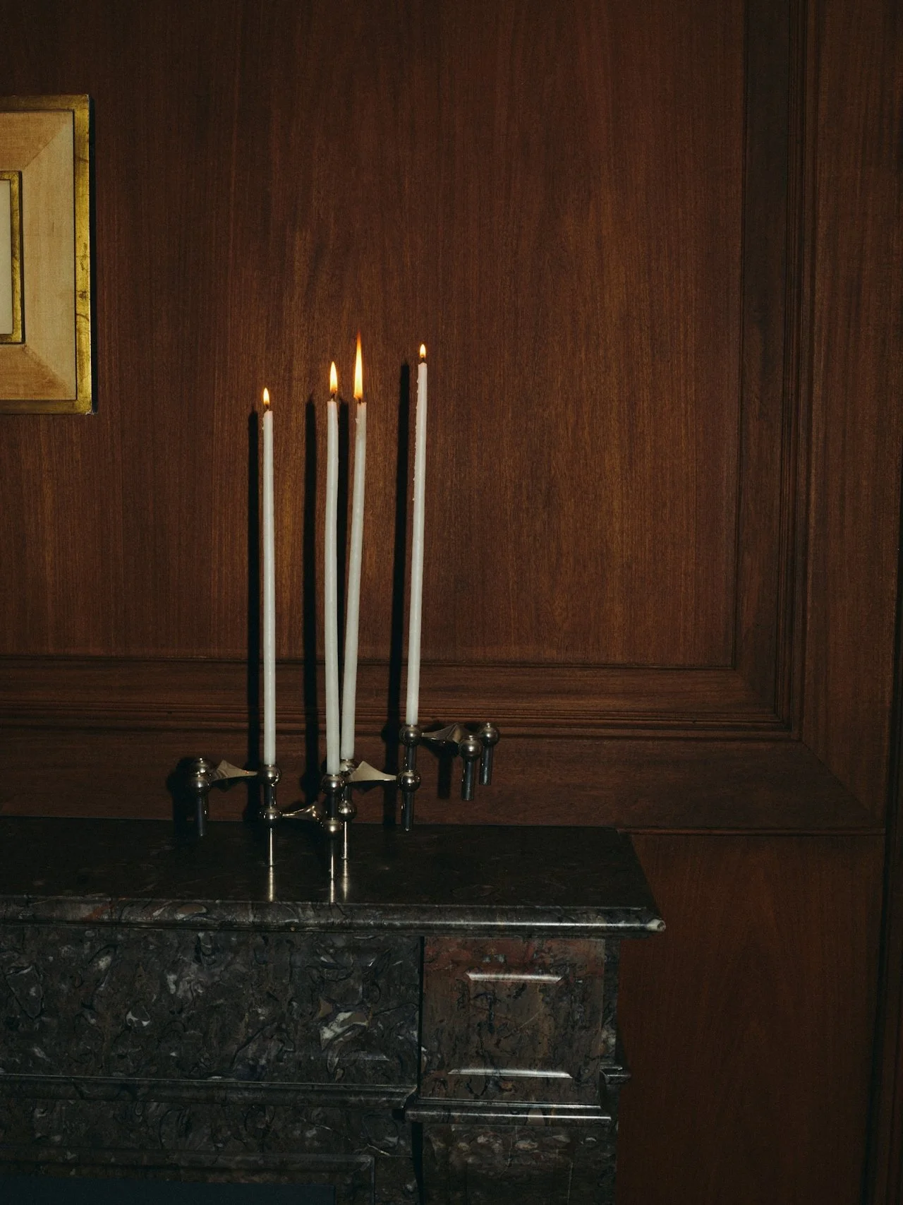 Four lit white candles in a candle holder on a dark marble table against a wooden paneled wall.