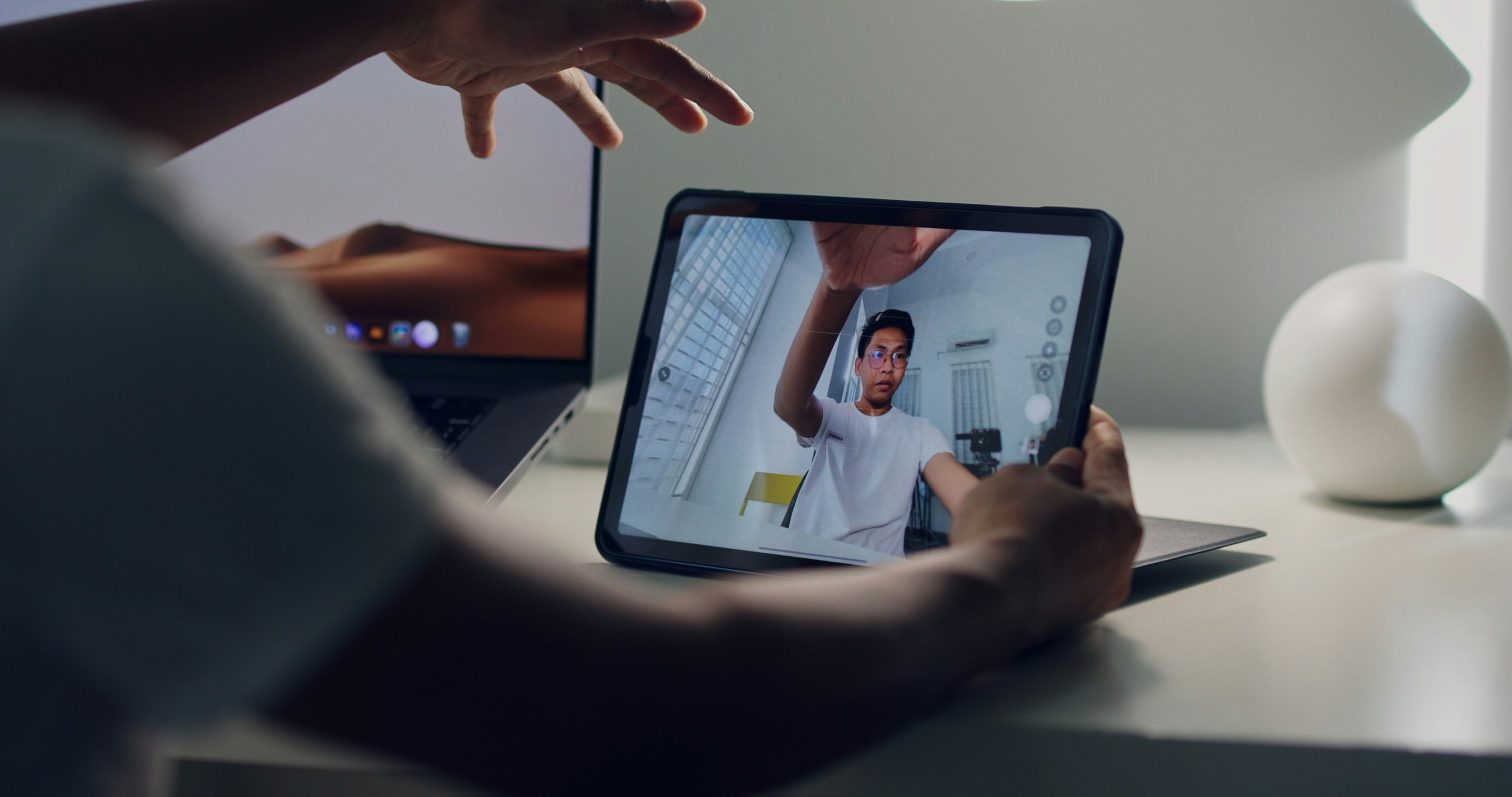 A person is taking a selfie with a tablet, capturing their reflection in the screen. The scene is set on a white desk, with a laptop and a white decorative object in the background.